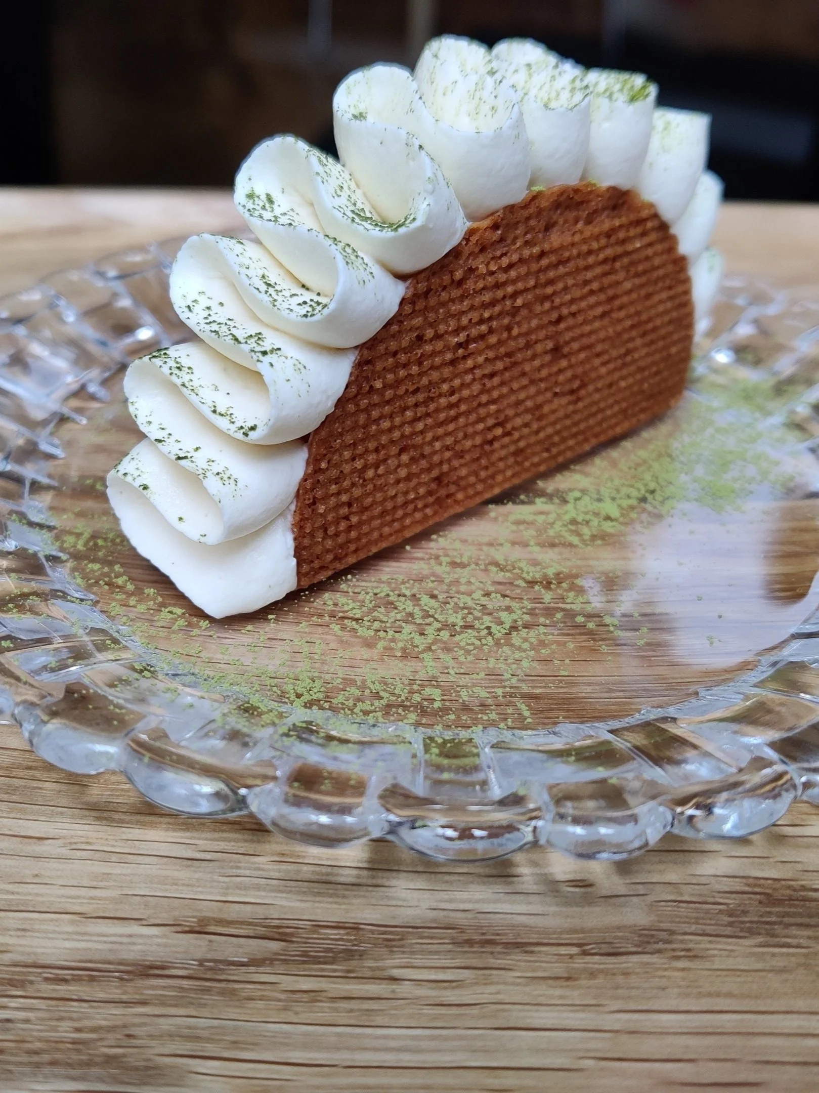 A piece of tiramisu dessert with layers of coffee-soaked sponge cake, topped with whipped cream and green powder, served on a glass plate.