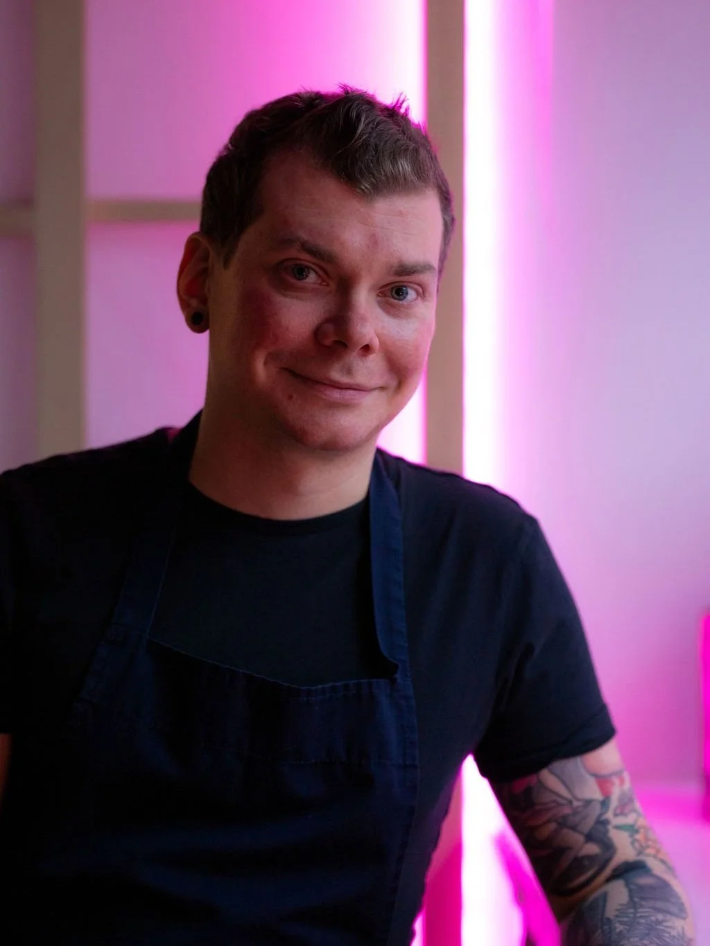 A young man with short brown hair, light skin, and tattoos on his arm, wearing a black T-shirt and apron, smiling in front of a pink neon-lit background.