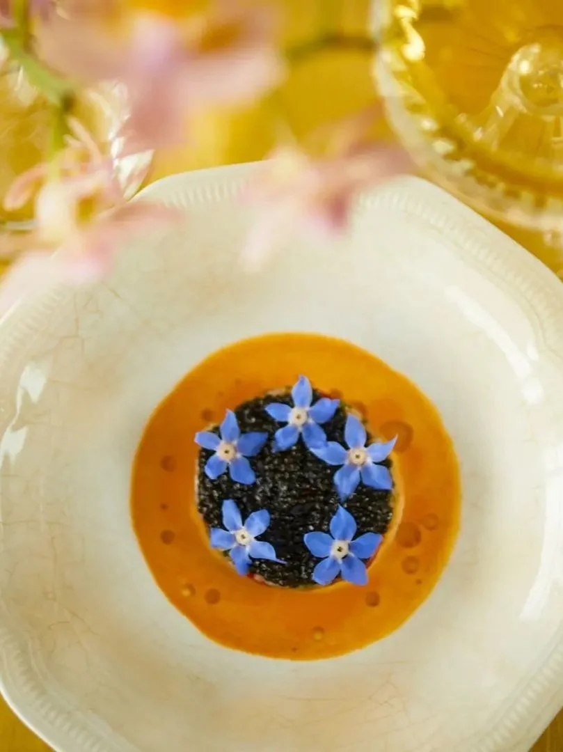 A white bowl with a dish topped with small blue flowers, surrounded by an orange sauce or puree, with some pink and yellow flowers and jars visible in the blurred background.