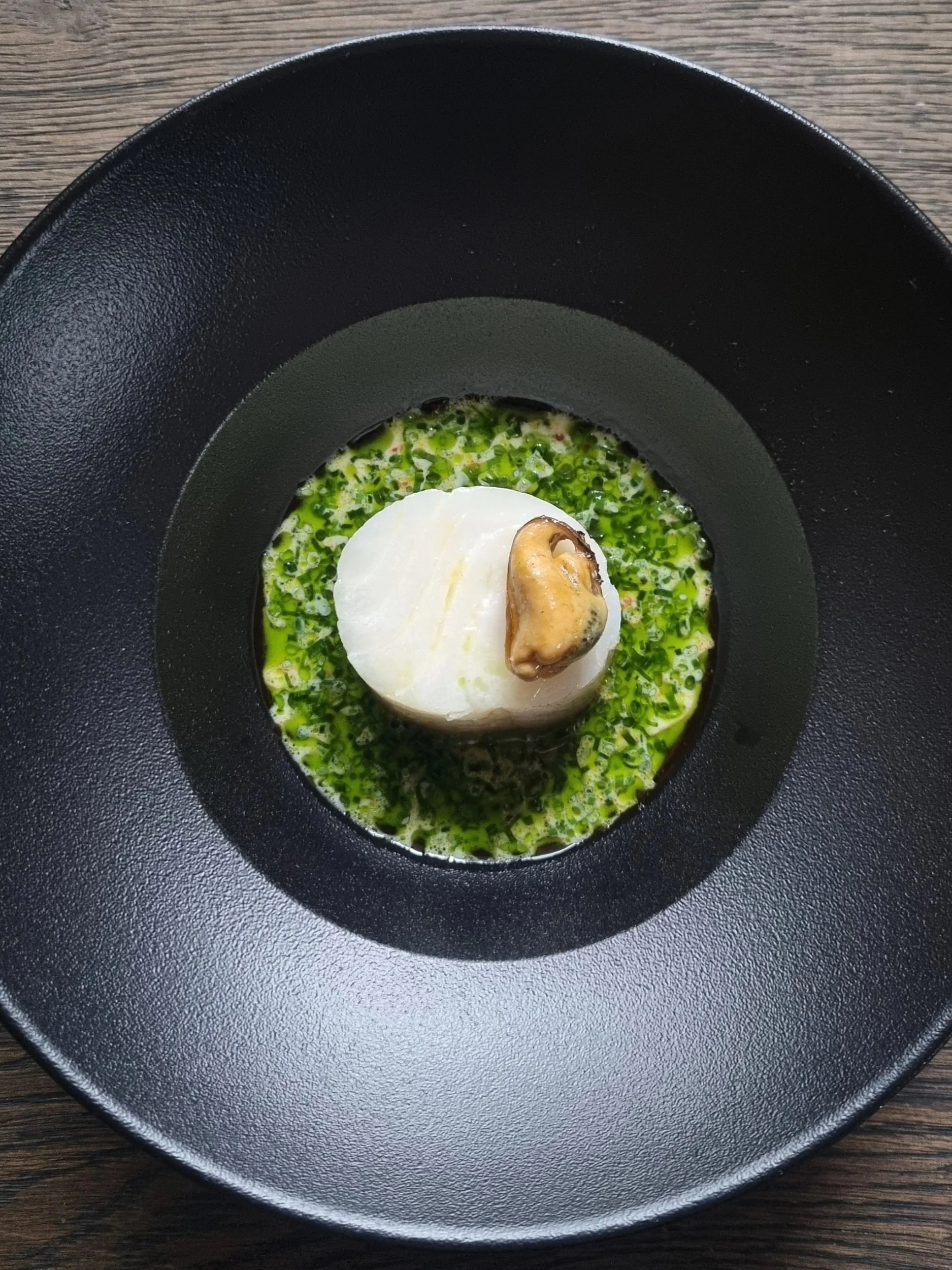A black bowl containing a piece of white fish topped with a mushroom, surrounded by a green herb sauce or oil, on a wooden table.