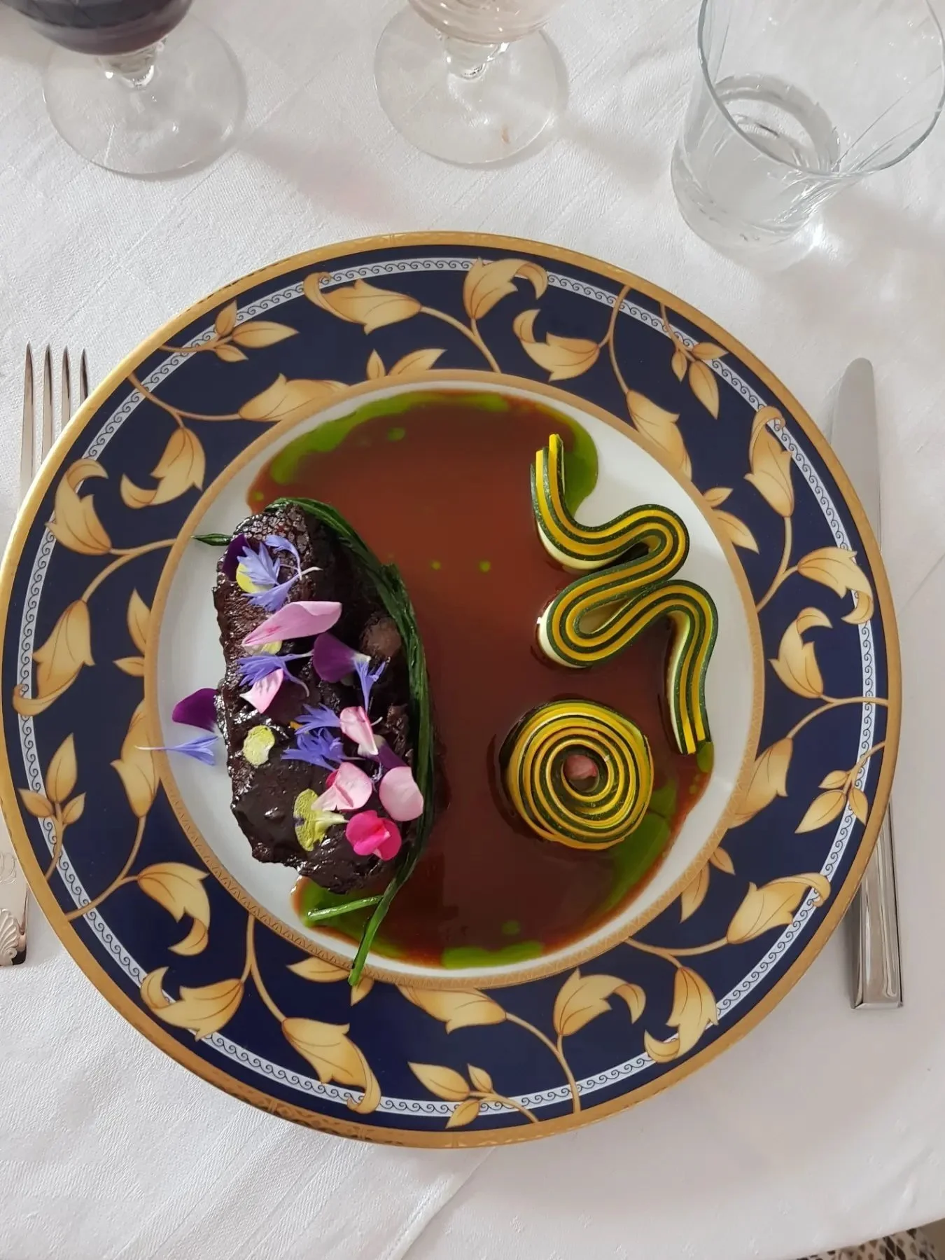 Elegant plated gourmet dish with a chocolate dessert, edible flowers, green sauce, and decorative green and yellow spirals on a white and blue ornate plate with gold accents, placed on a white tablecloth with cutlery and glasses nearby.