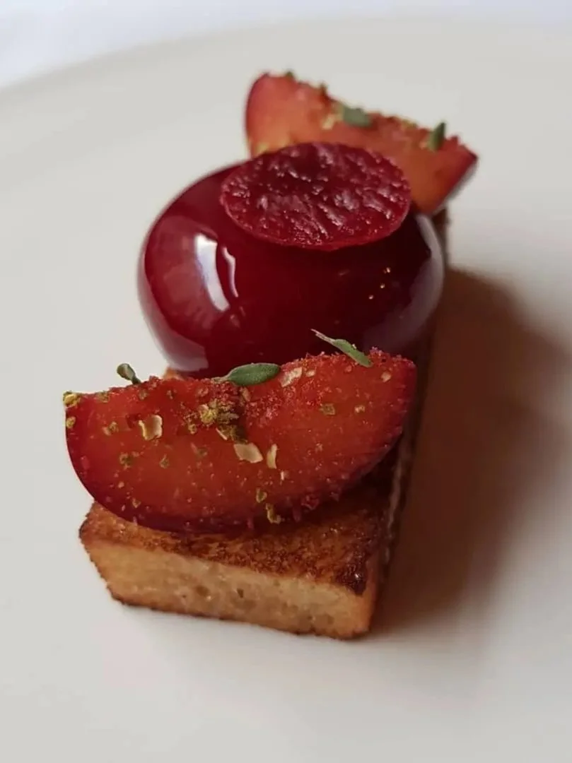 A plated dessert featuring a square piece of toasted bread topped with a cherry, sliced strawberries, and garnished with small green herbs.