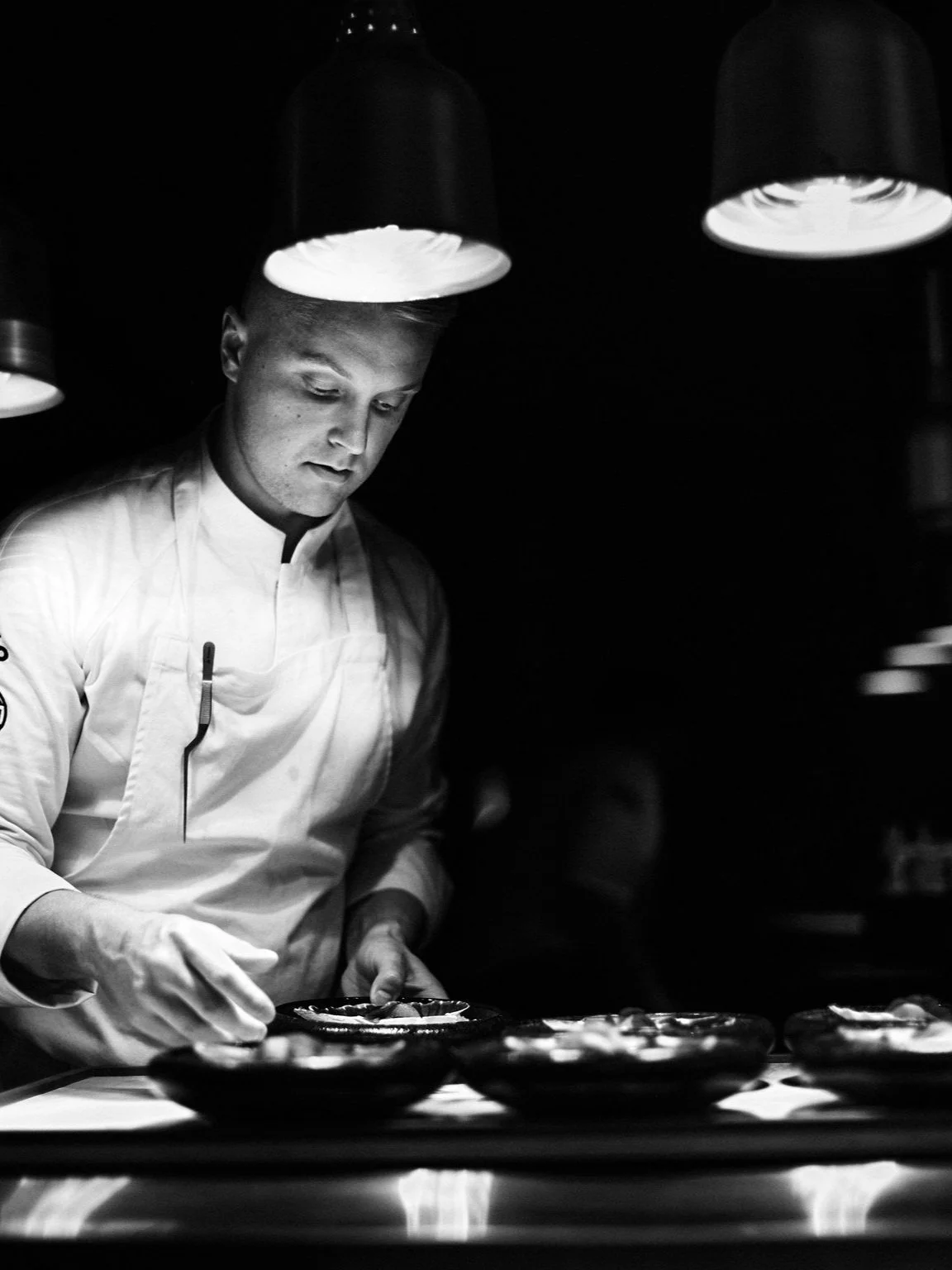 Black and white photo of a chef in a professional kitchen, carefully preparing dishes under overhead lighting.
