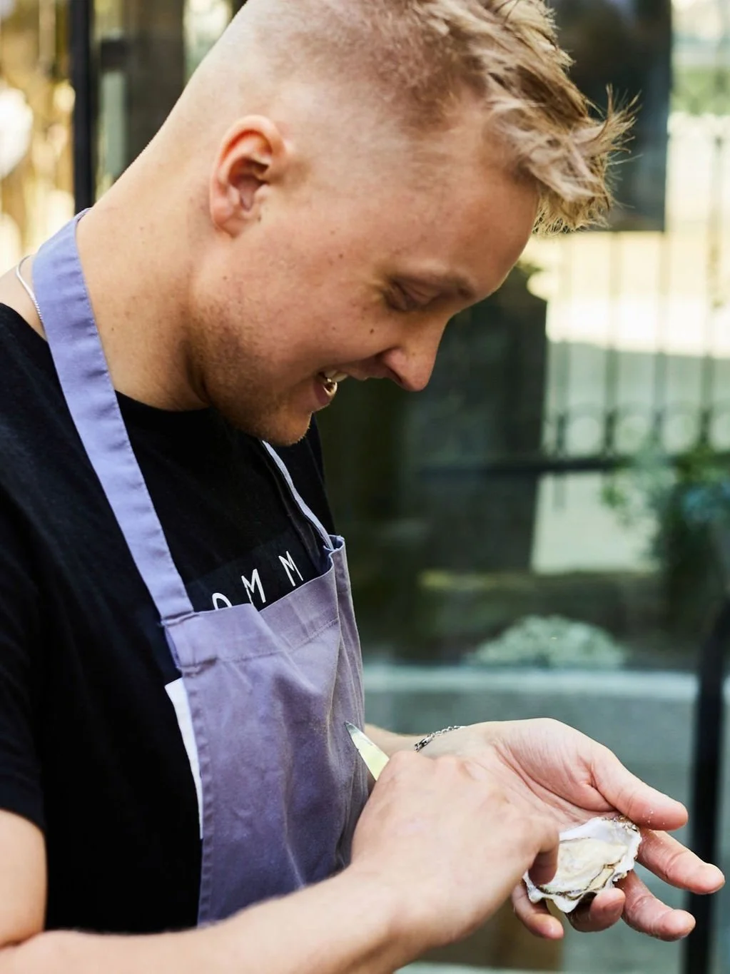 A young man wearing a gray apron opening an oyster outdoors.