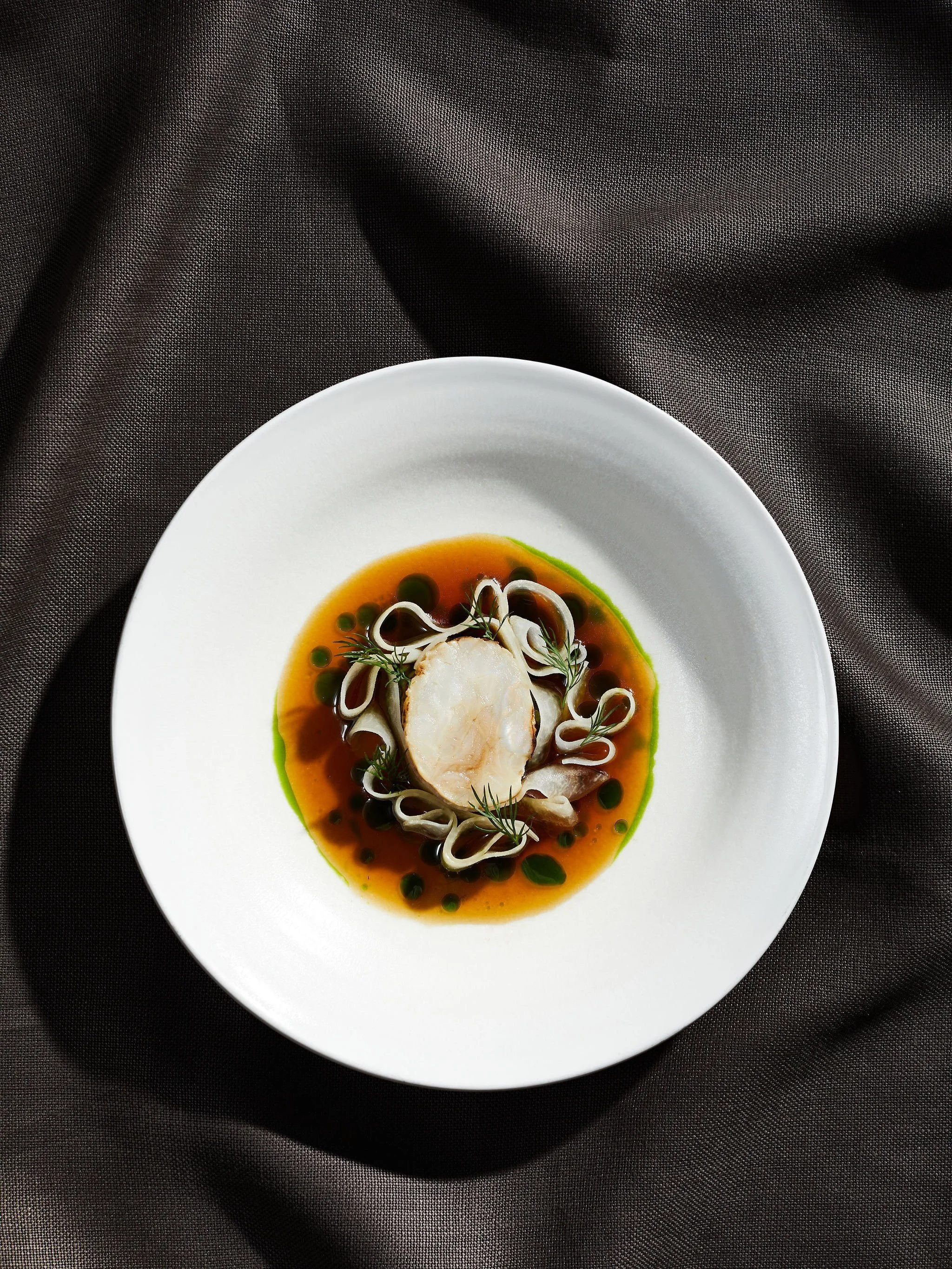 White plate with cooked fish topped with herbs, surrounded by a brown sauce with green droplets, garnished with small herbs, on a dark fabric background.