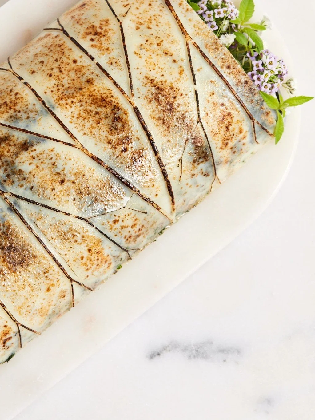 A rectangular cake coated with white icing, decorated with chocolate drizzle and cacao powder, garnished with purple flowers and green leaves on a white plate.