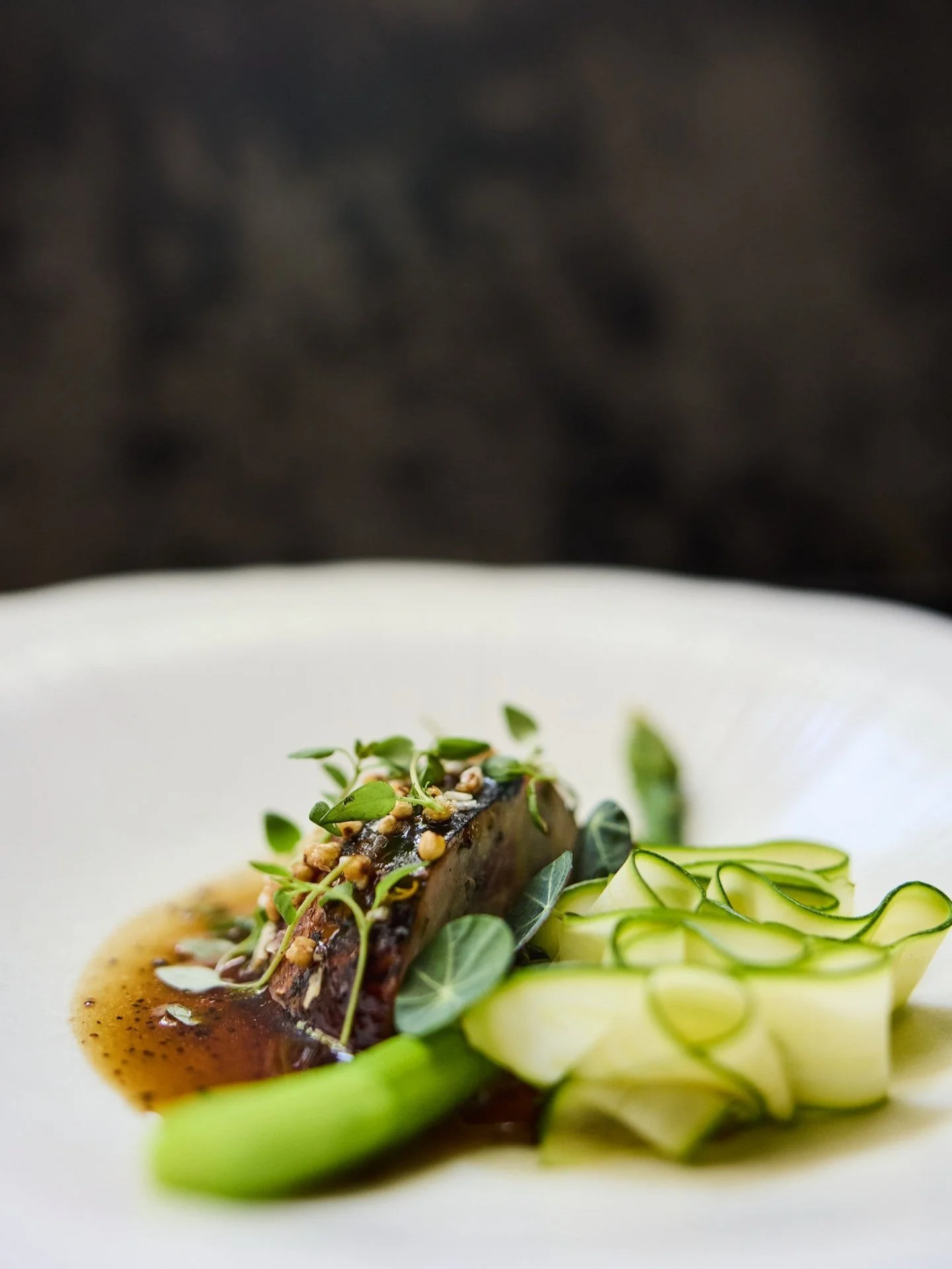 A gourmet dish with a slice of meat topped with microgreens and sauce, served with thinly sliced zucchini ribbons and a green vegetable on a white plate.