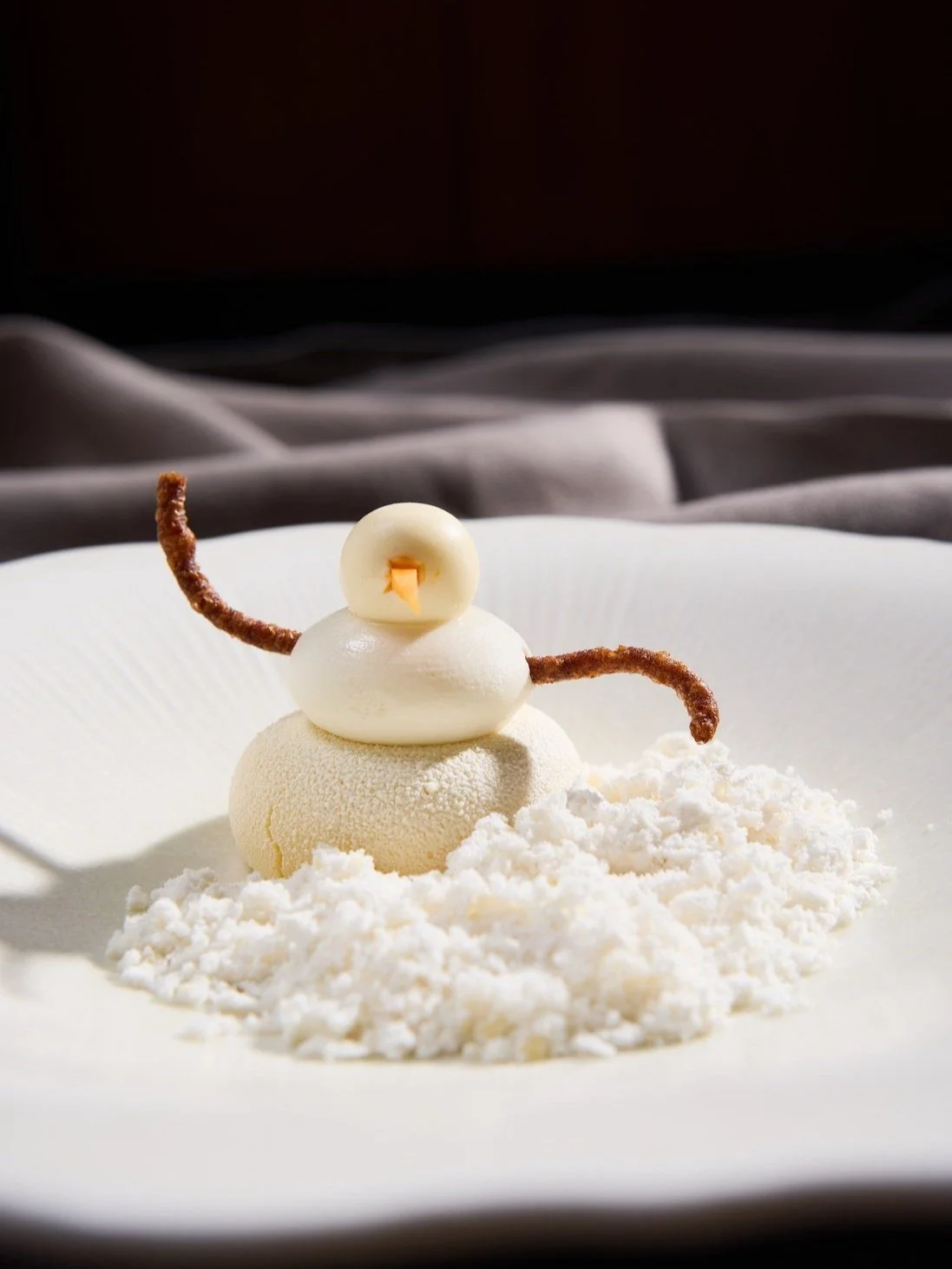 A snowman-like dessert made of white mousse or ice cream, with a round white chocolate piece for the head, small orange decoration for the beak, and pretzel sticks for the arms, all on a bed of white powdered sugar on a white plate.