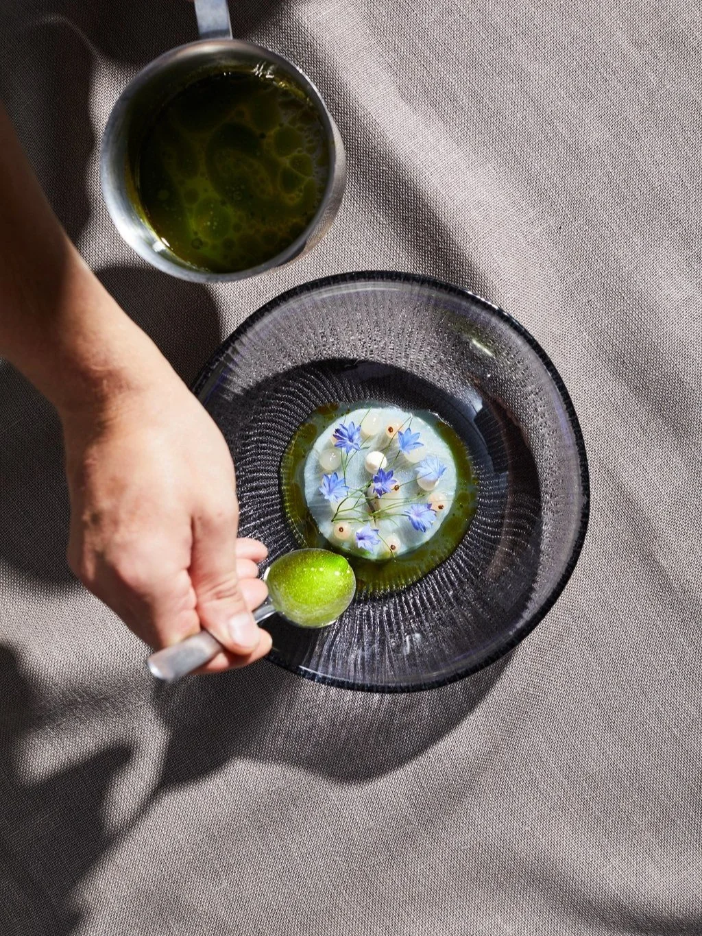 A hand holding a lemon on a spoon over a glass bowl containing blue flowers and white spheres in a green liquid, with a mug of green liquid nearby on a beige tablecloth.