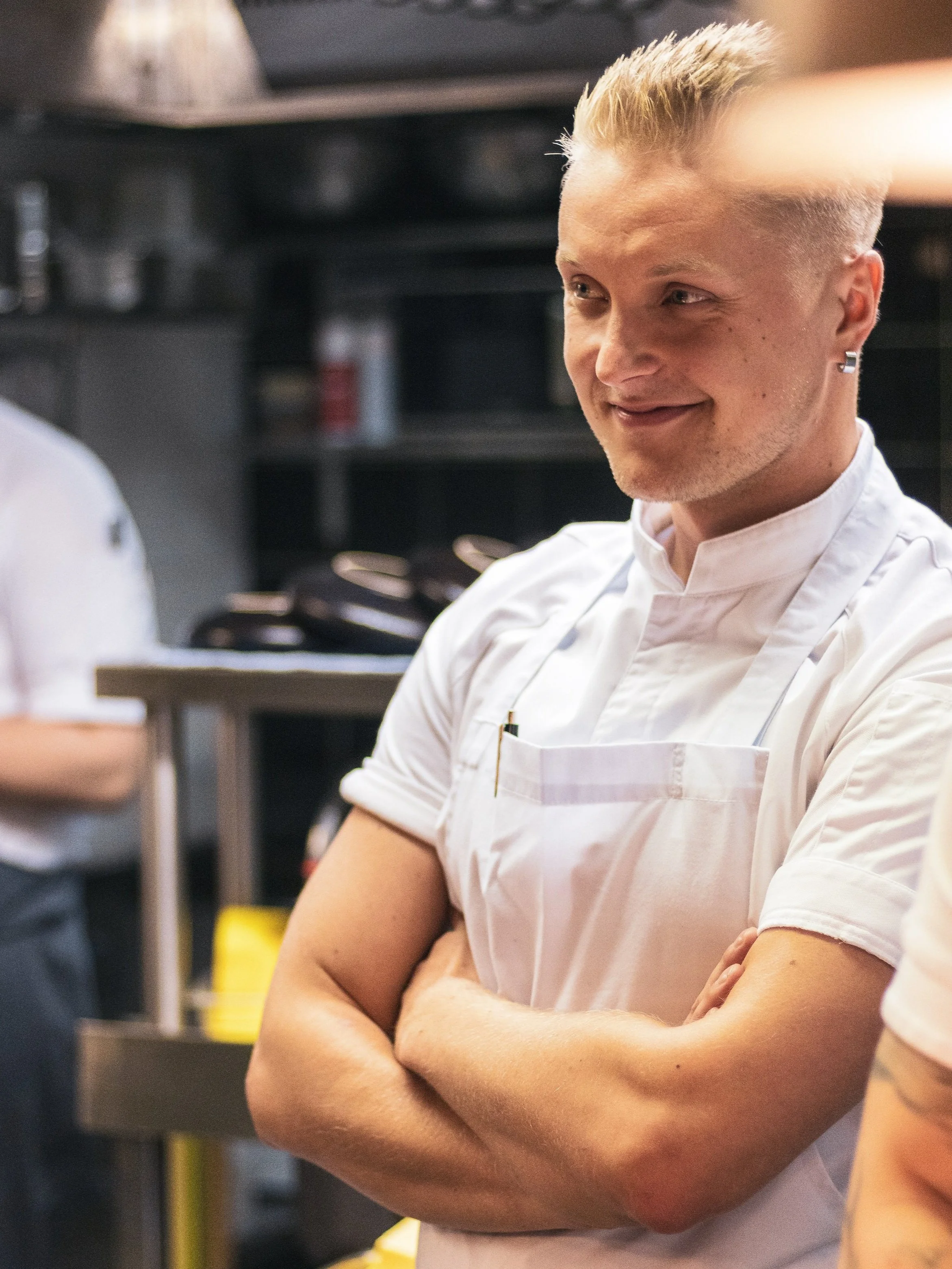 A young man in a white chef's coat and apron, smiling with arms crossed in a kitchen.