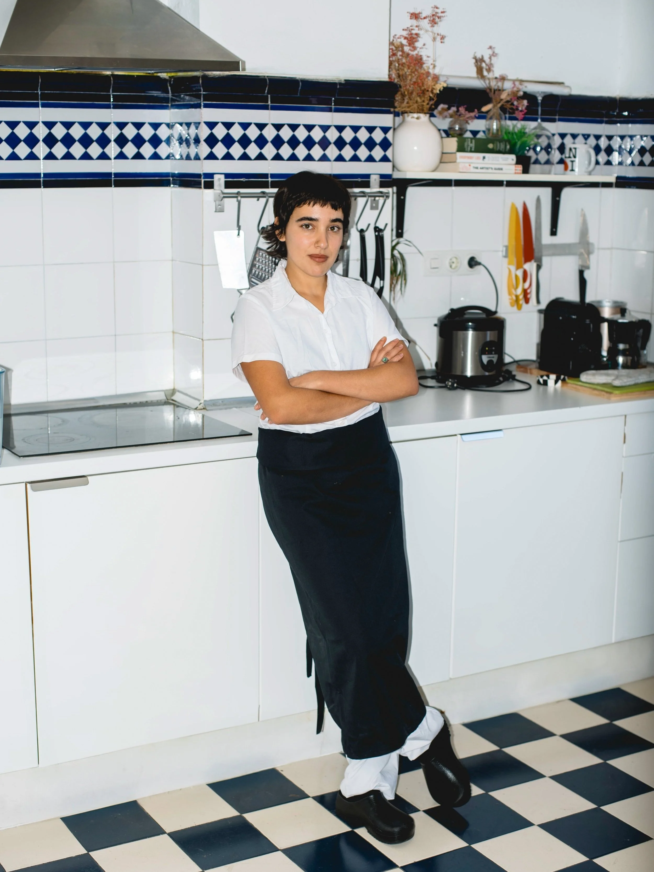 Woman standing with arms crossed in a kitchen, wearing a white shirt, black apron, and black shoes, with kitchen utensils and appliances in the background.
