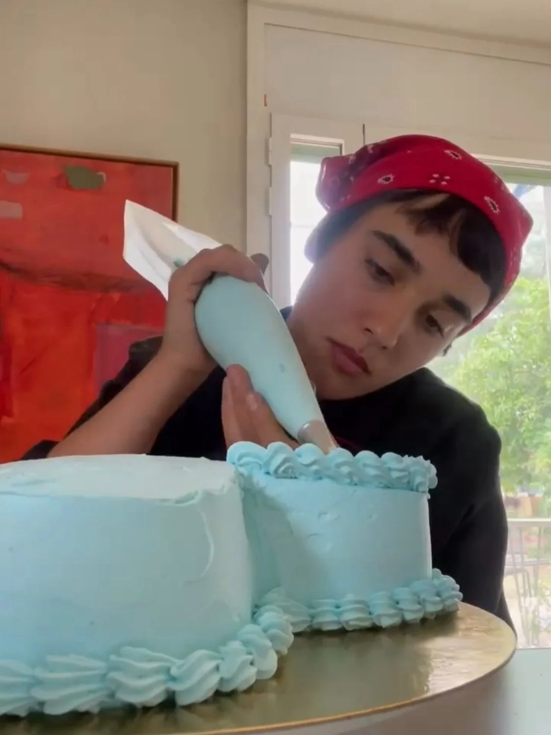 A young person wearing a red bandana decorates a light blue cake with white frosting using a piping bag.