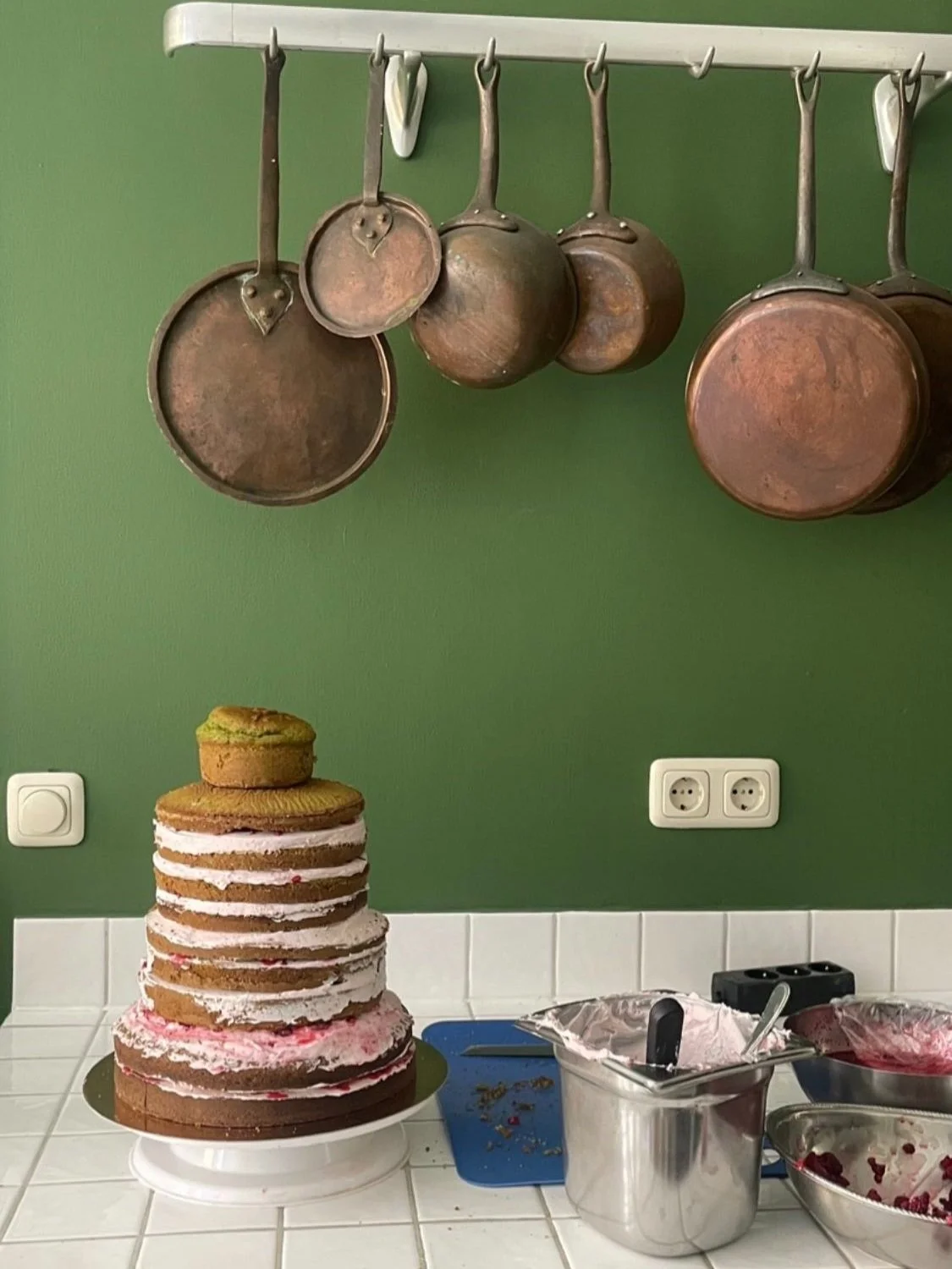 A partially assembled layered cake with pink and white filling, topped with a single cookie, on a white cake stand. Behind it, a blue cutting board and assorted bowls with cake decorating tools and remnants are visible on a white tiled countertop. Co