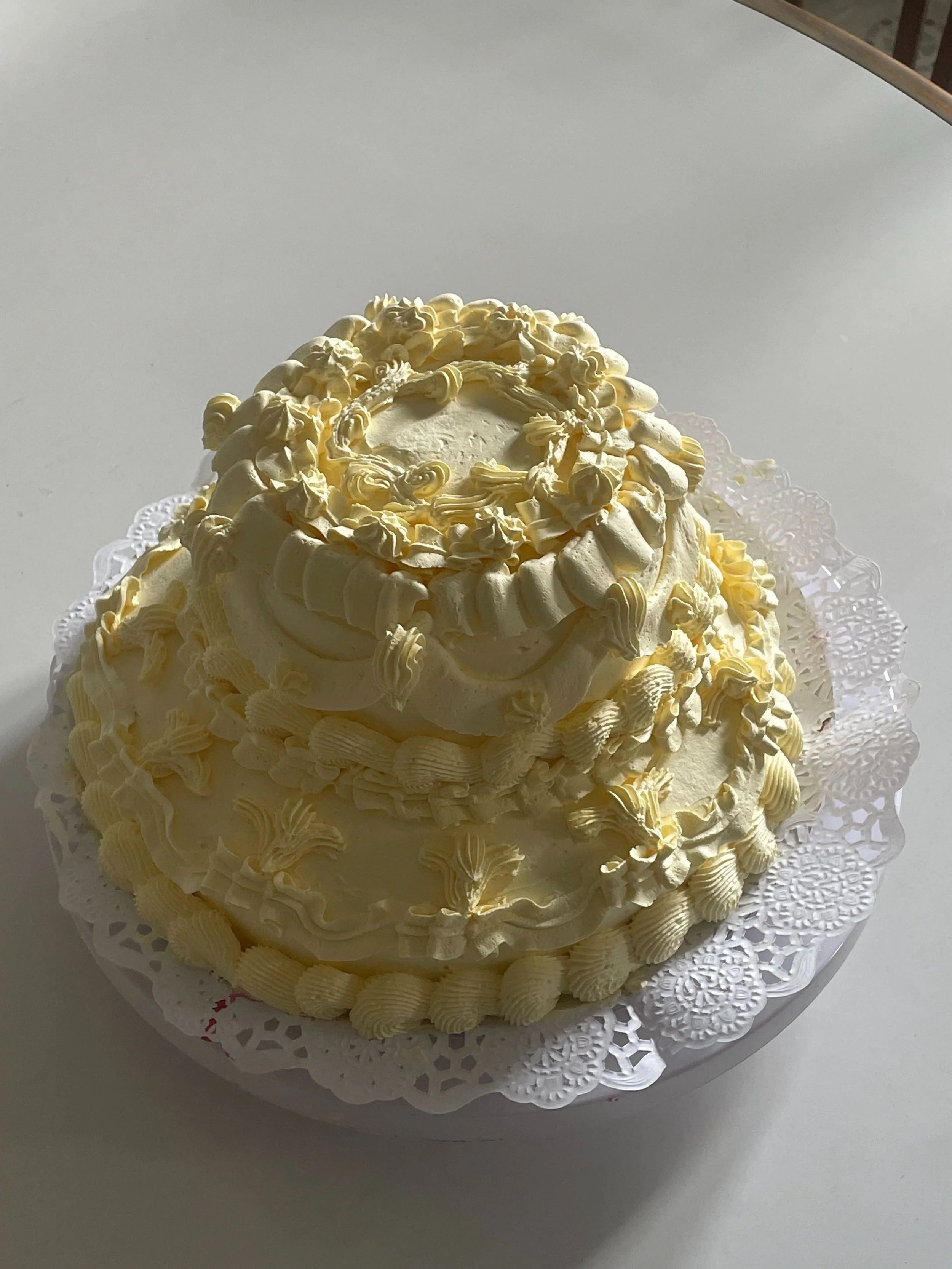 A white, tiered cake decorated with cream-colored frosting, piped in floral and scroll designs, sitting on a decorative paper doily.