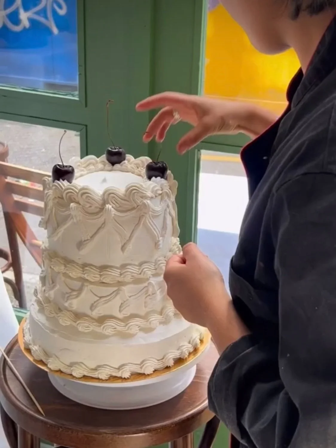 A person is decorating a large white birthday cake with cherries on top and white decorative frosting.