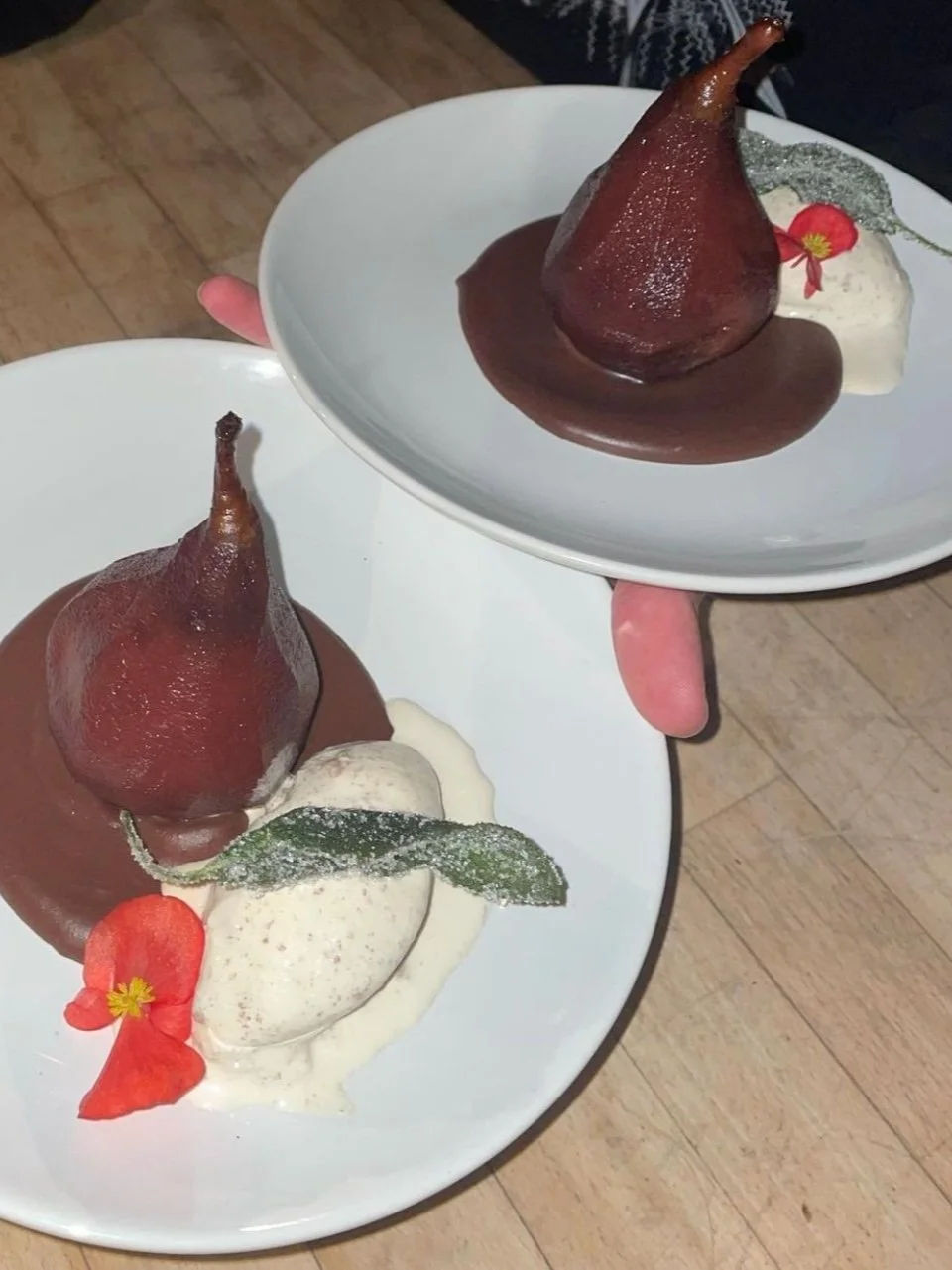 Two plates with poached pears, ice cream, and garnishes, served with chocolate sauce. The pears are decorative and topped with edible flowers and leaves.
