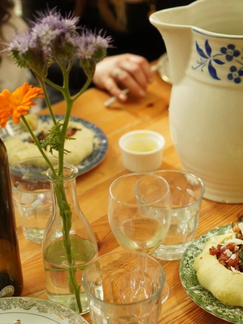 A dining table set with a small bouquet of purple and orange flowers in a glass bottle, several glasses of water, a pitcher with a blue floral design, a white condiment dish, and a plate with a glazed pastry or bread topped with a mixture of chopped