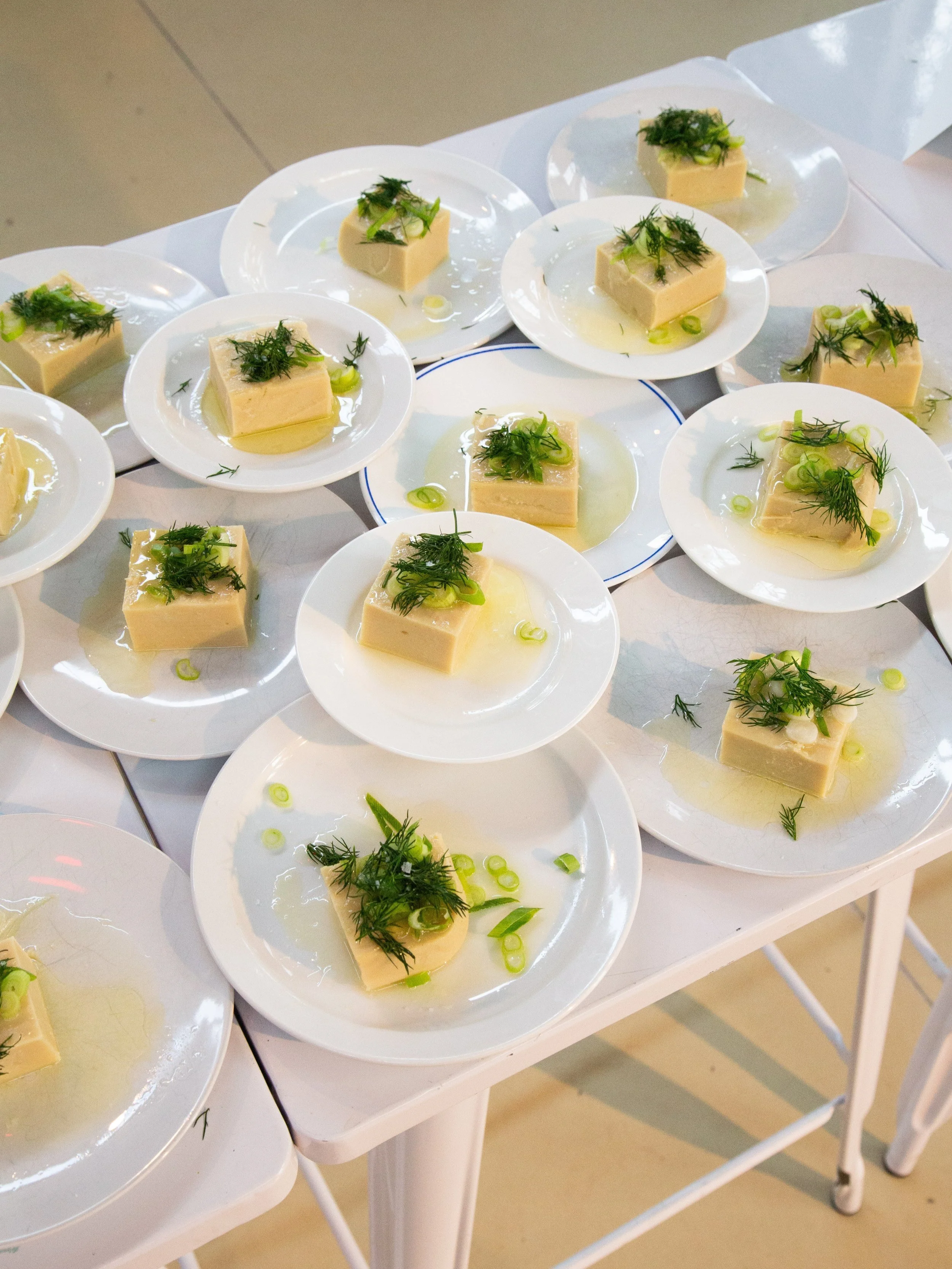 Small white plates with cubed tofu garnished with green herbs and sliced green onions, drizzled with oil, arranged on a white table.