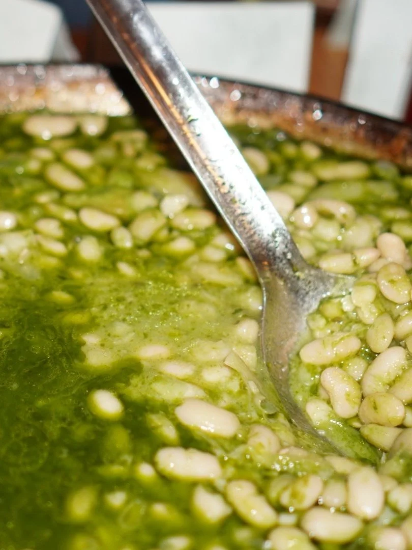 A large metal spoon stirring cooked white beans in a green sauce or broth.