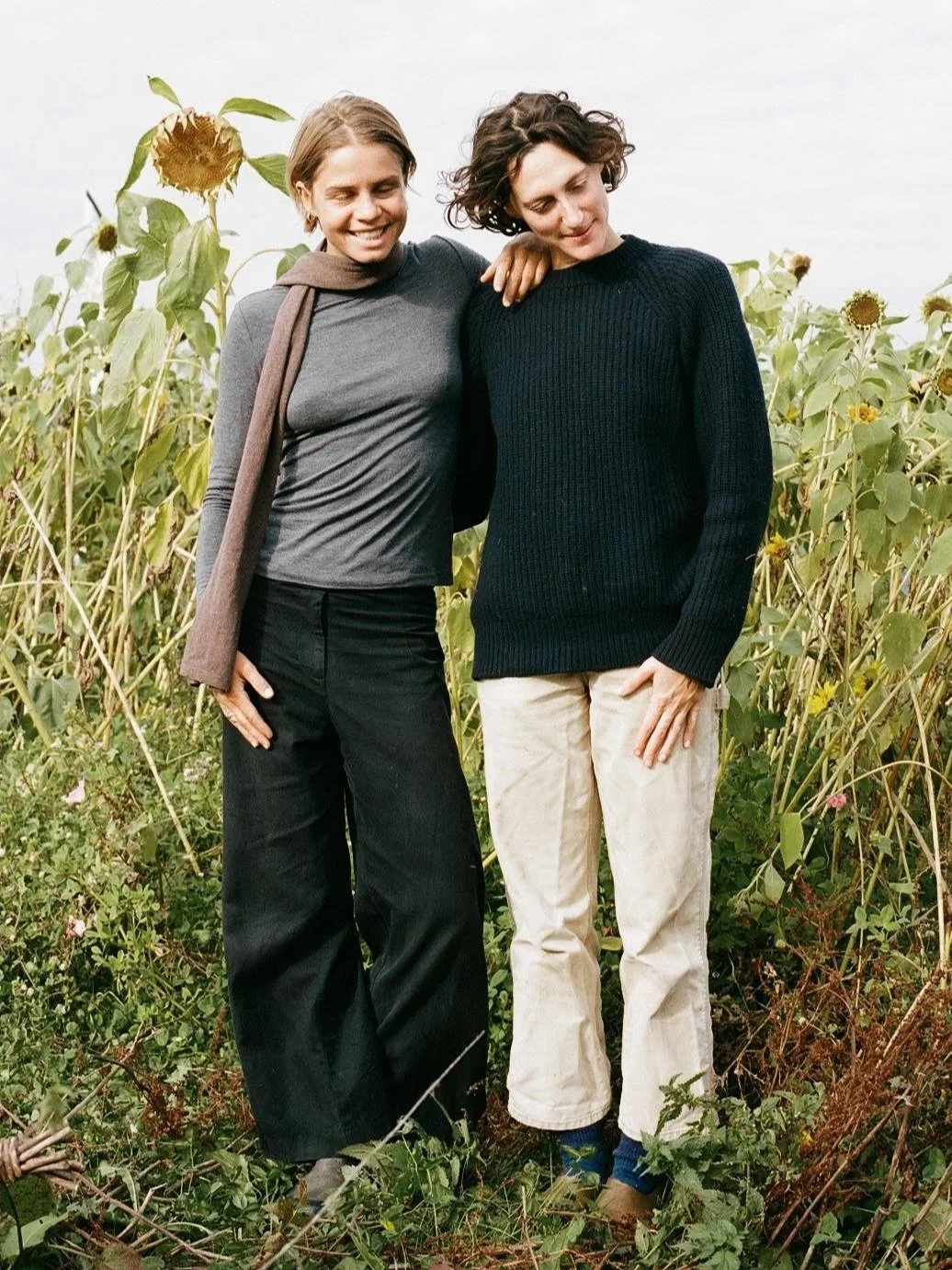 Two women standing in a sunflower field, smiling and leaning on each other, with their eyes closed.
