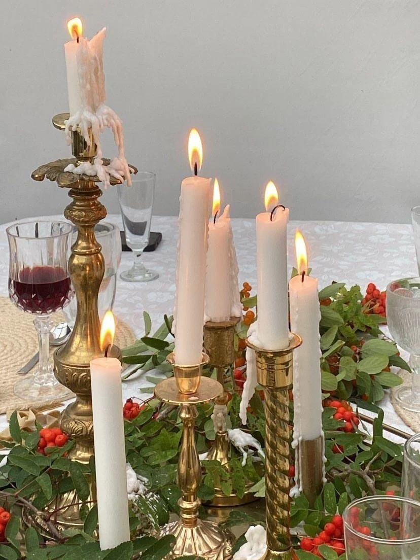 A dining table decorated with lit candles, some melted, in brass candlesticks, and a centerpiece of greenery with red berries, set for a meal. A glass of red wine is visible, along with empty glasses and tableware.