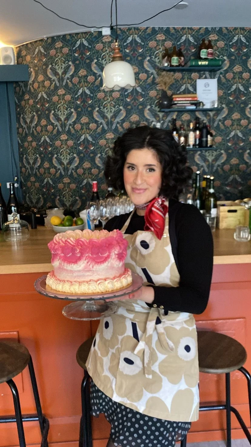 A woman with dark curly hair wearing a black shirt, patterned apron, and red bandana around her neck is smiling and holding a decorated pink and white birthday cake on a glass cake stand in a cozy, decorated bar or café setting.