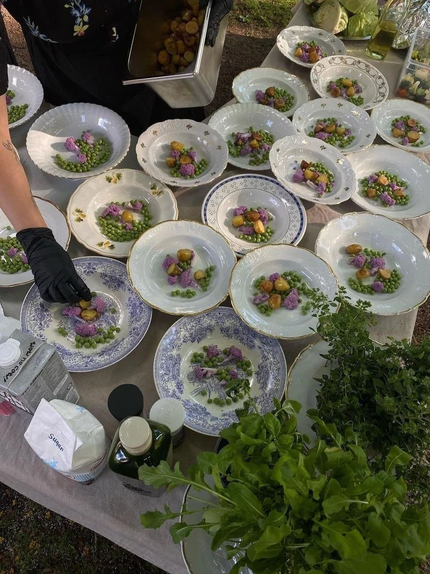 Several decorative plates on a table, each with small servings of food including green peas, purple cauliflower, and yellow potatoes, with a person wearing black gloves preparing one of the dishes.