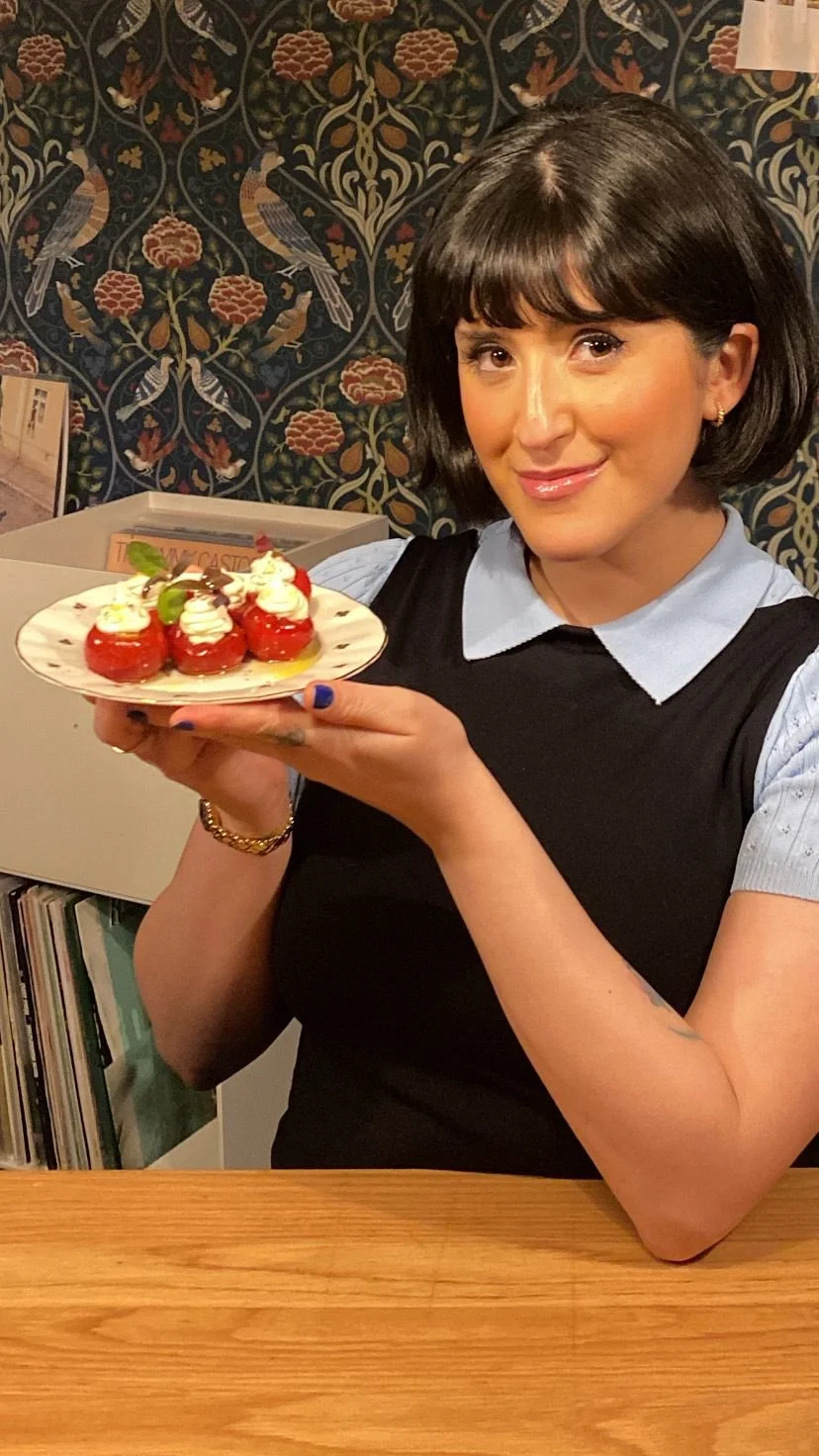 A woman with dark hair and wearing a black sleeveless top with a light blue shirt underneath, holding a small plate of decorated pastries with strawberries and whipped cream. She is smiling at the camera. Behind her is a patterned wallpaper with bird