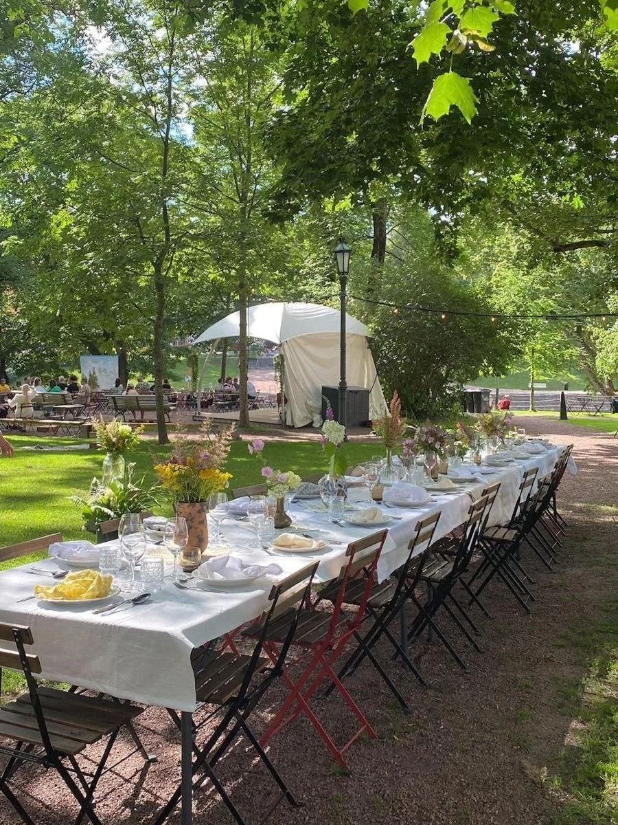 Long outdoor dining table set with white tablecloth, glassware, and floral centerpieces in a lush green park with trees, picnic benches, and a white tent in the background.