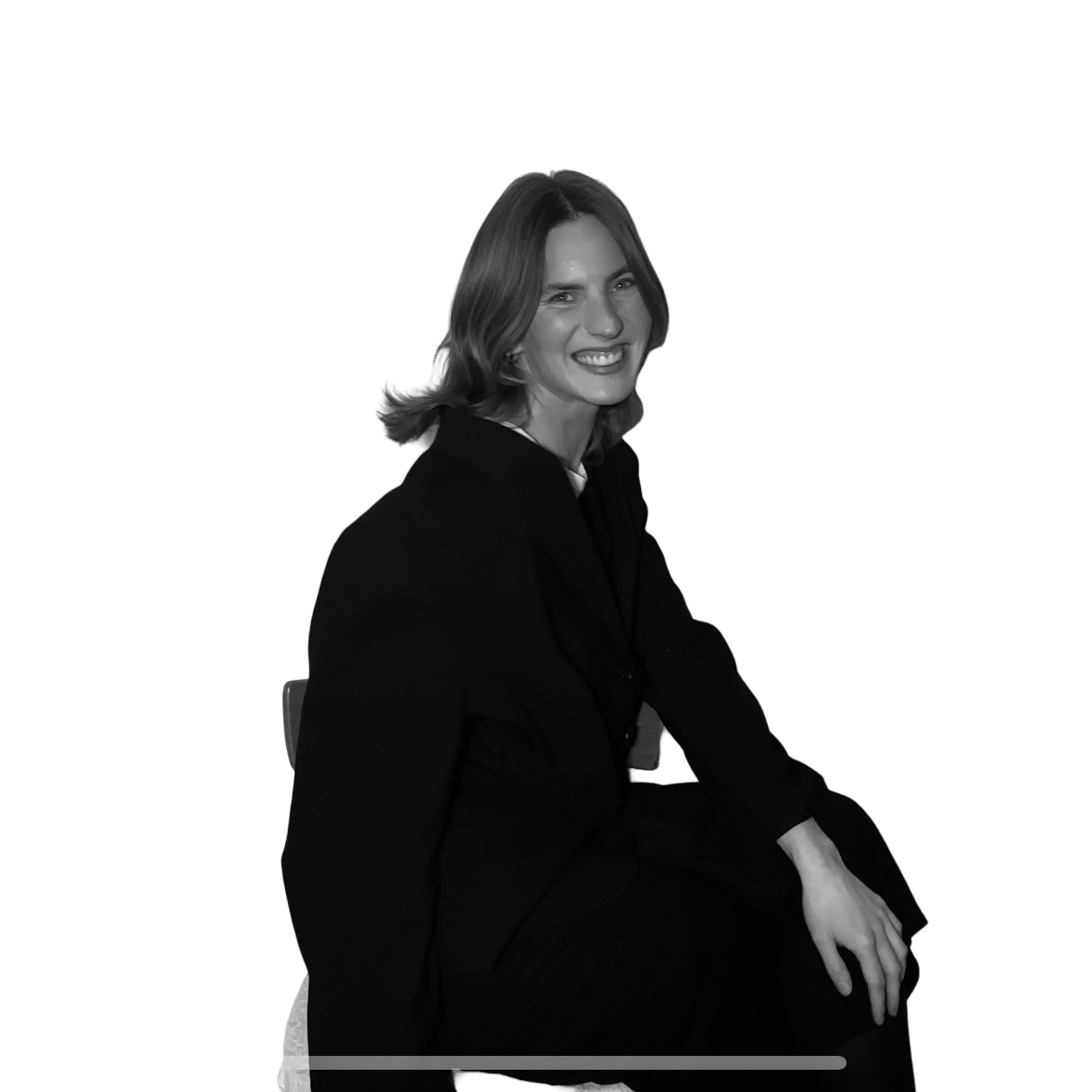 Black and white image of a woman with shoulder-length hair, wearing a dark blazer, sitting on a chair, smiling at the camera.
