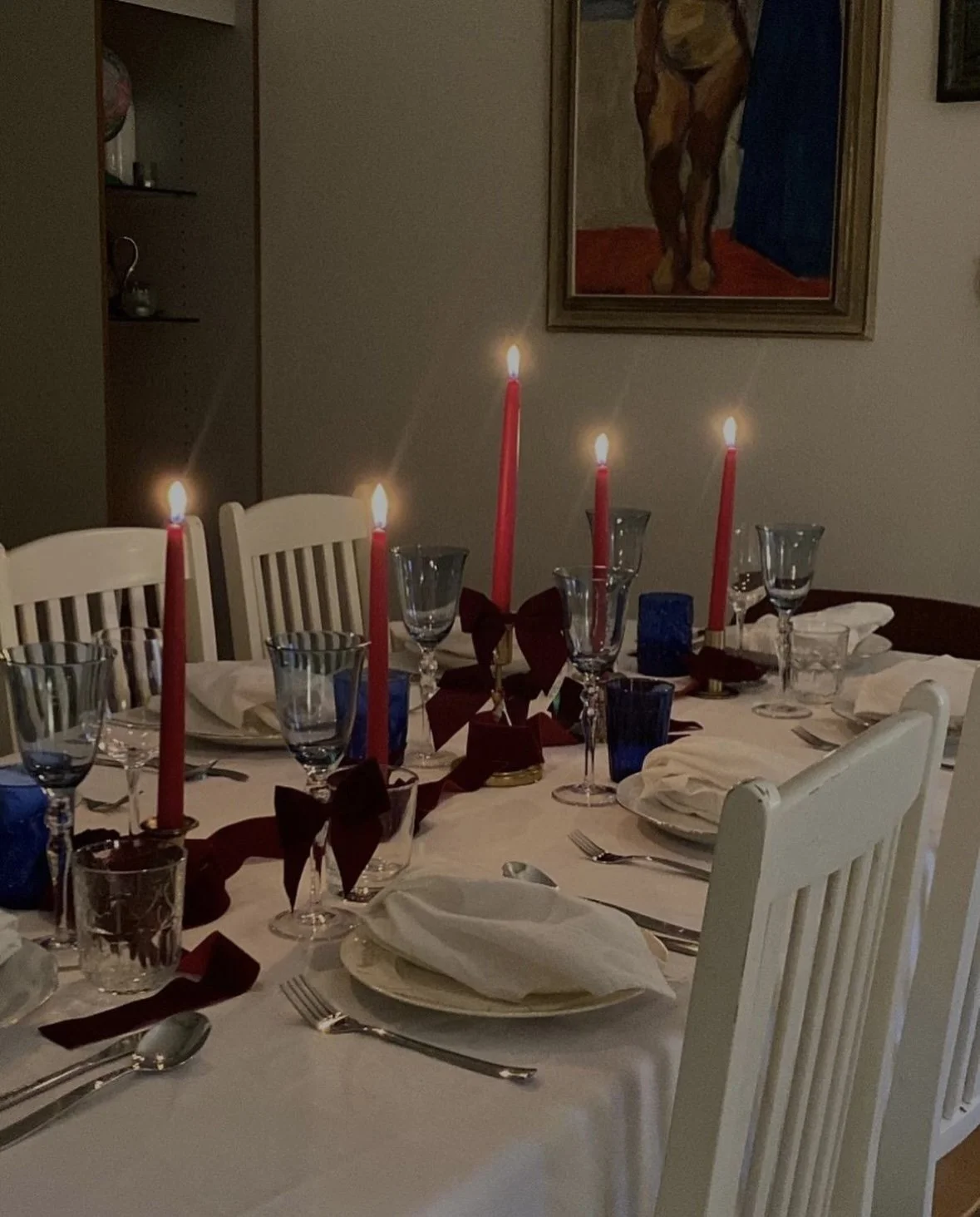 A dining table set for a formal dinner with white linens, multiple glassware, and cutlery. Five red candles are lit in the center, adorned with bows. There are folded white napkins on each plate, and a framed painting of a horse hangs on the wall in