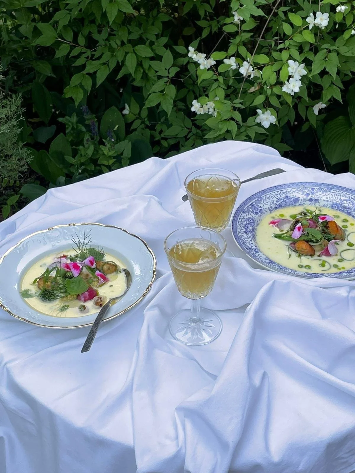 Elegant outdoor table setting with two plates of white soup garnished with herbs and edible flowers, two glasses of a light-colored beverage, and a white tablecloth amidst green foliage and white flowers.