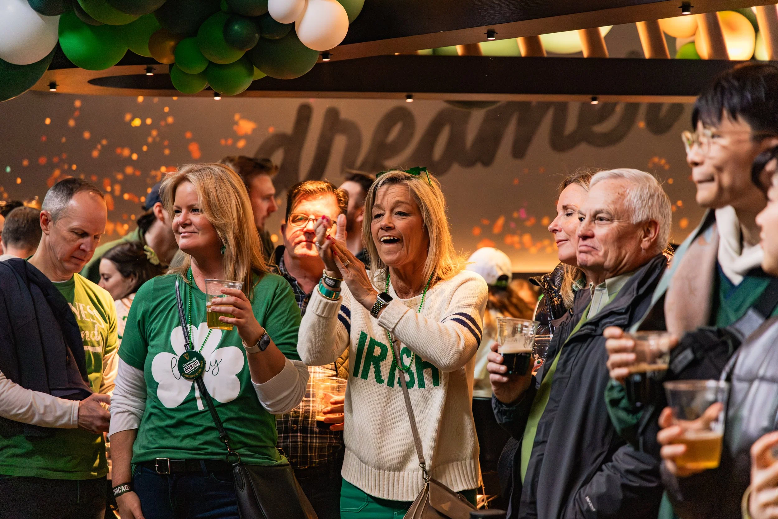 People gathered at a celebration event, wearing green and Irish-themed clothing, with some holding drinks and cheering.