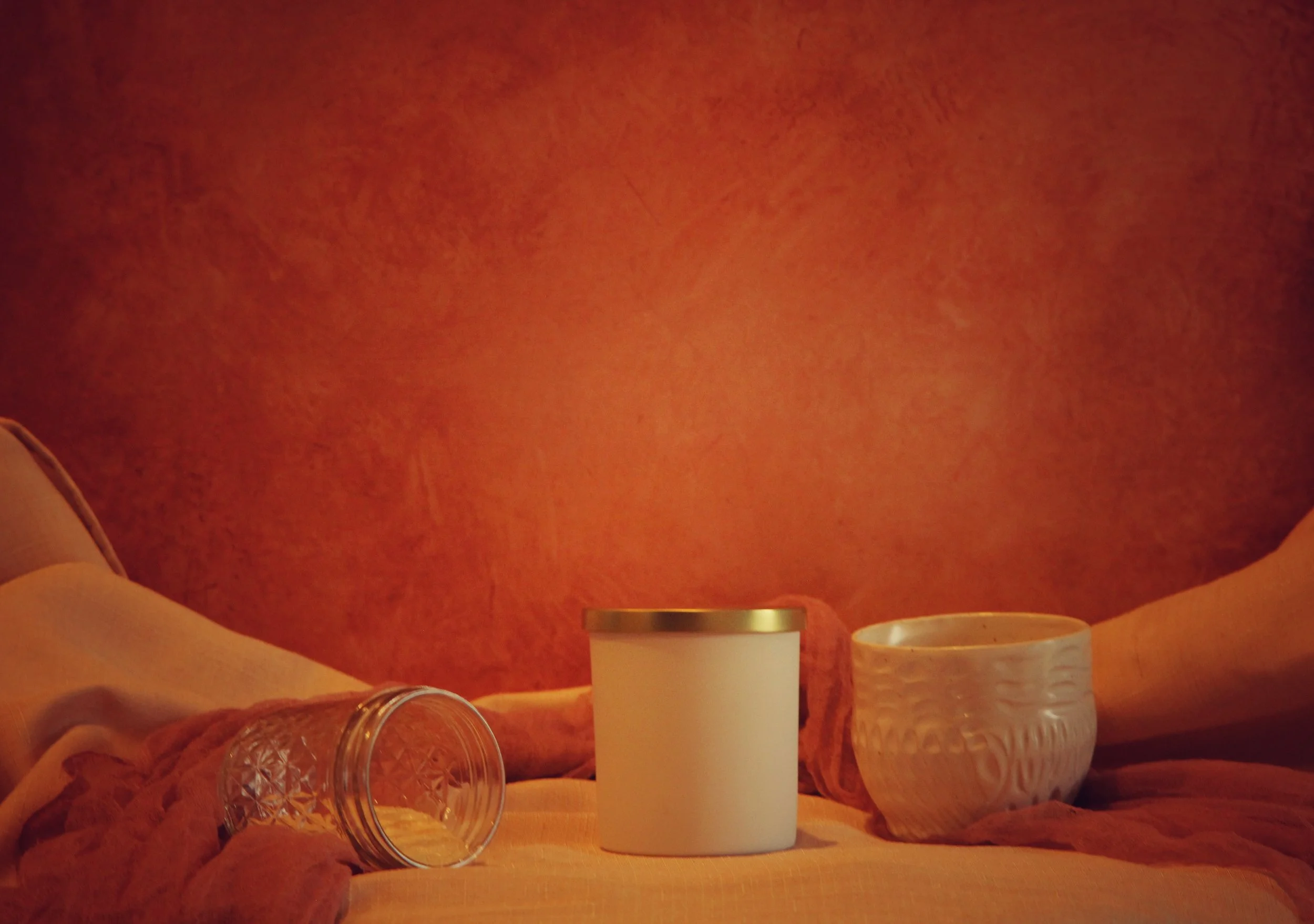 A still life scene with a glass tumbler lying on a soft surface, a white container with a gold lid, and a textured white bowl against a warm orange background.