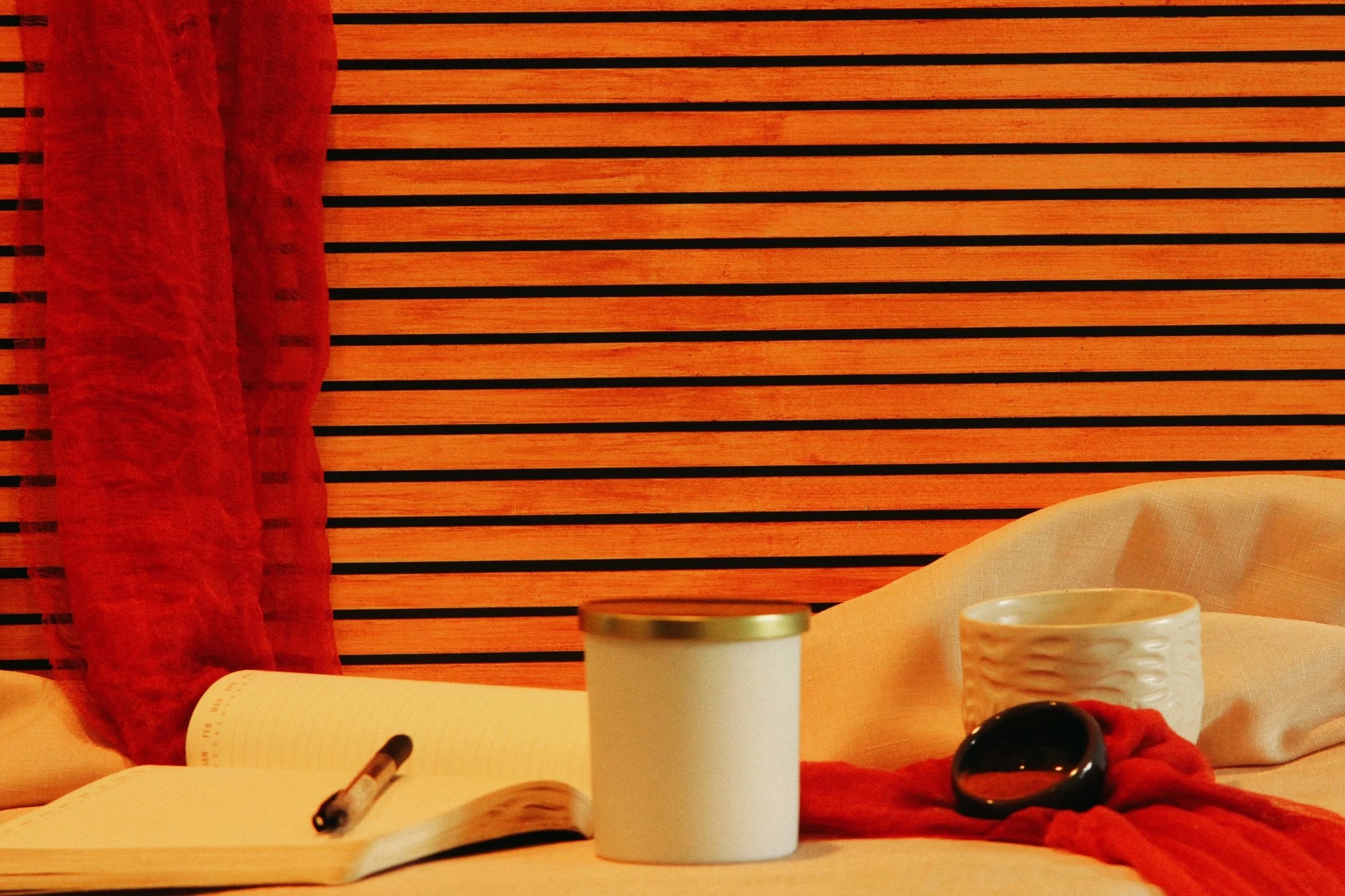 A bed with an open notebook and pen, a white tumbler, a ceramic cup, a red scarf, and a black ashtray, set against a wooden-paneled headboard with a red curtain on the left side.