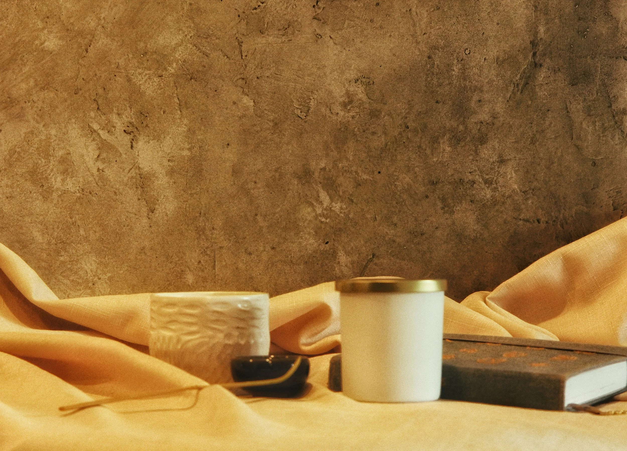 A beige fabric with a textured ceramic mug, a white paper cup with a metallic lid, a black device, and a closed book with a checkered pattern on its cover, all placed against a rough, textured wall.