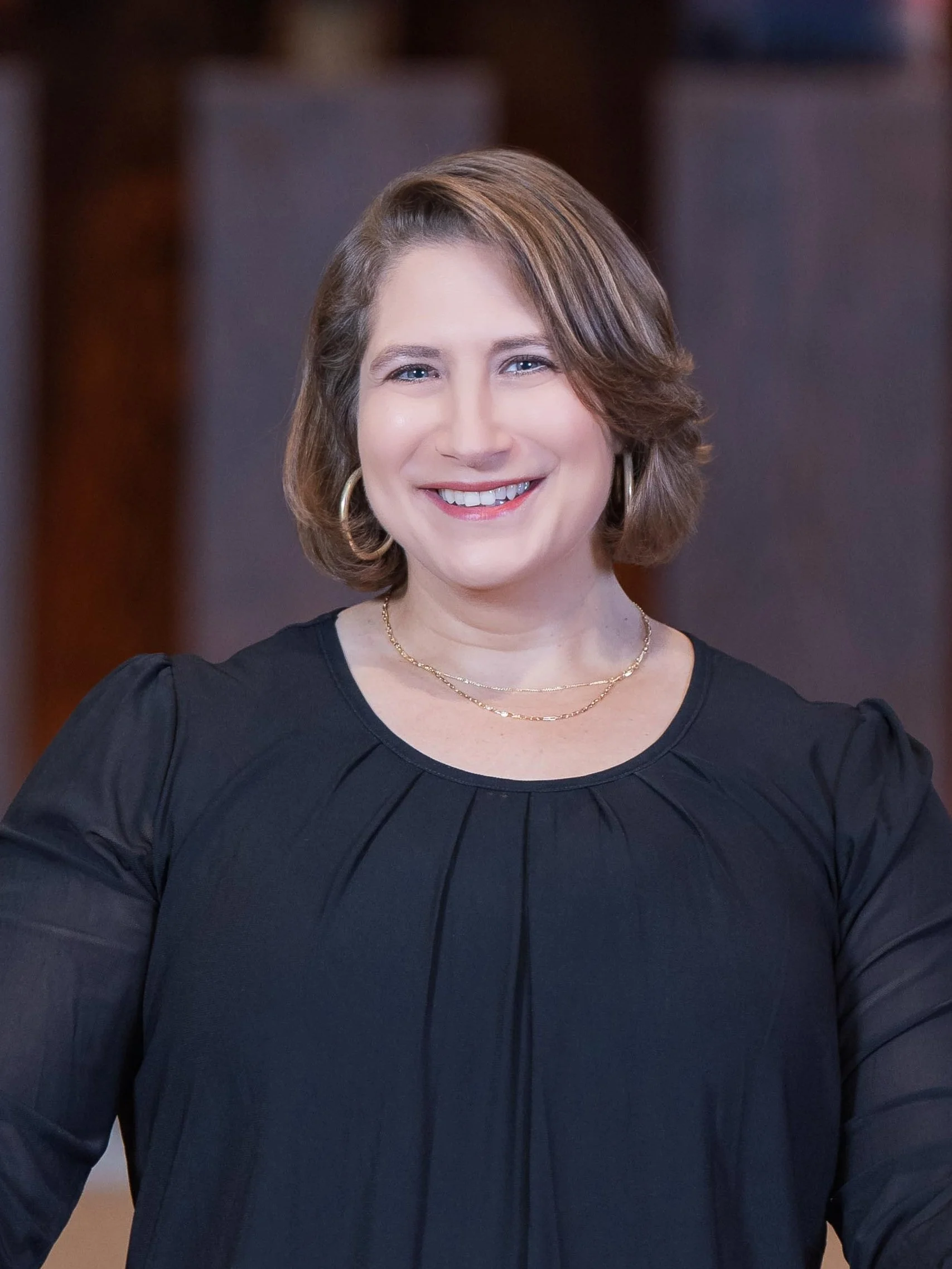 Senior Rabbi Elizabeth Sacks standing in front of a sand-colored brick wall, wearing a black shirt.