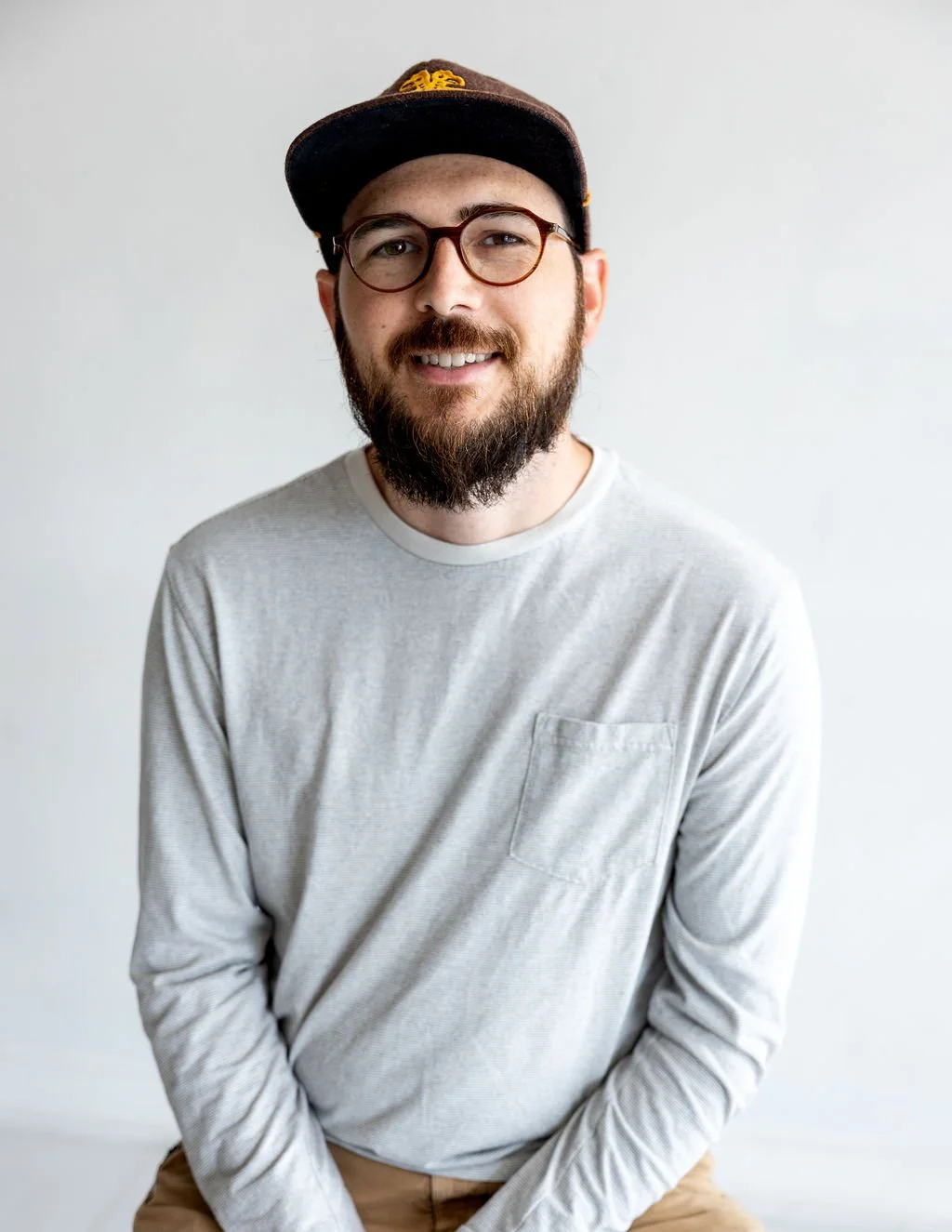 A smiling man with a beard, glasses, wearing a baseball cap, white long-sleeve shirt, and khaki pants, sitting against a plain white wall.