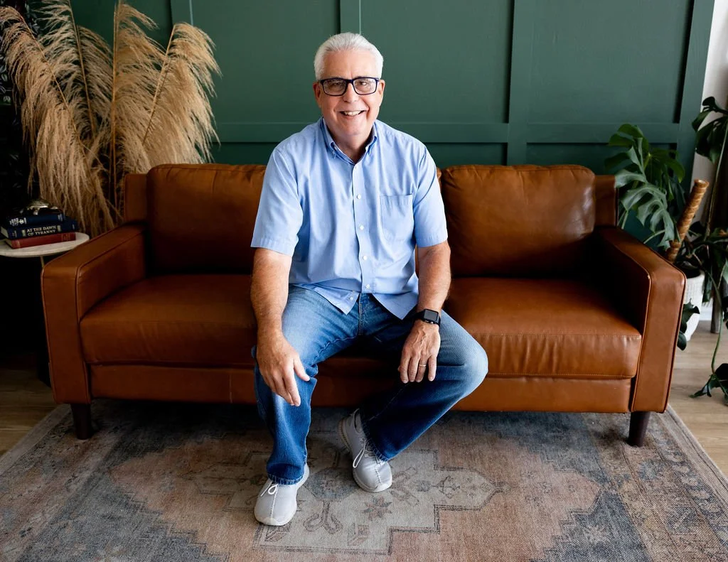 A smiling older man with glasses, wearing a light blue short-sleeve button-up shirt, blue jeans, and white sneakers, sitting on a brown leather sofa in a room with green walls and various plants.