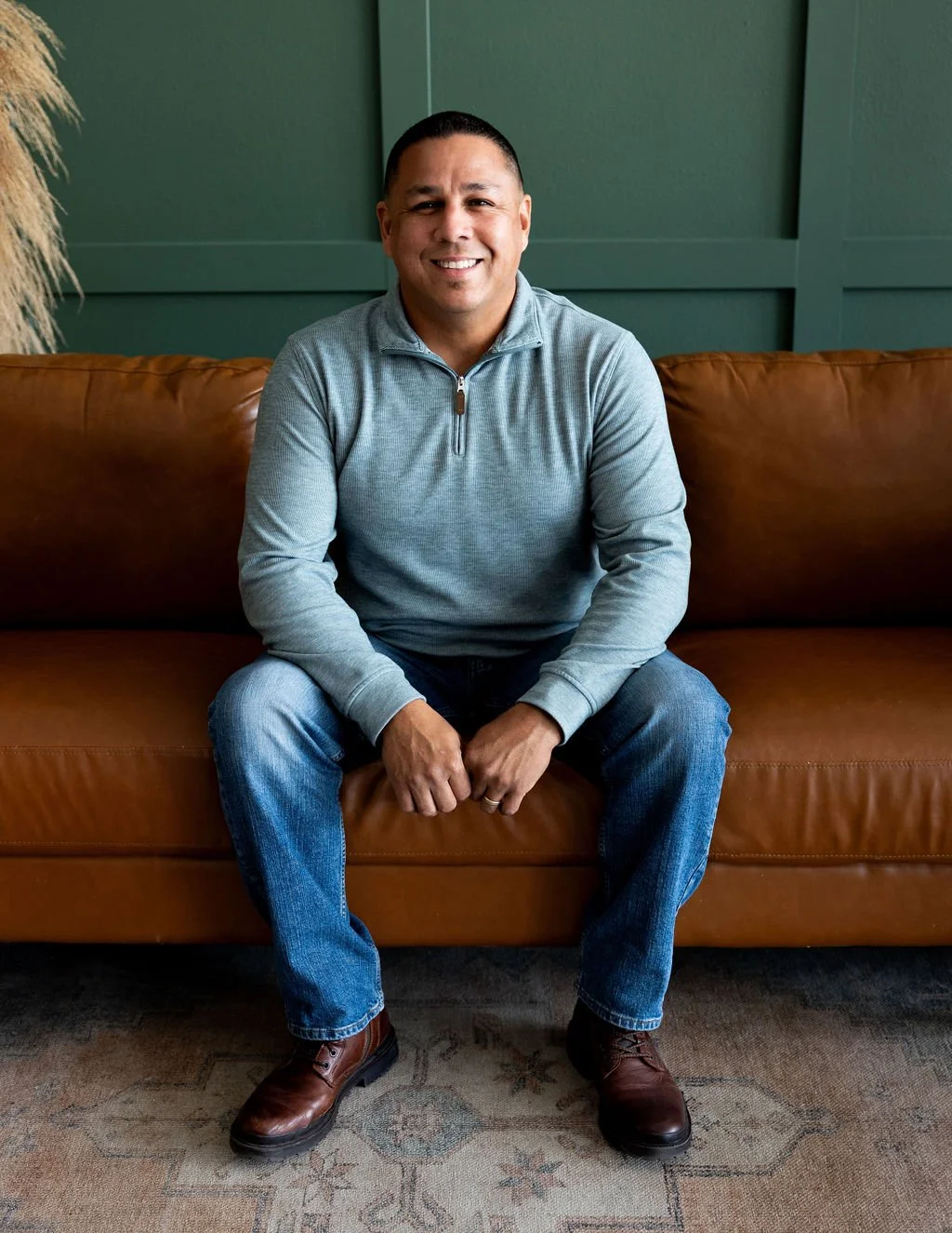 A man sitting on a brown leather couch, smiling, wearing a light gray zip-up sweatshirt, blue jeans, and brown boots, with a green wall background.