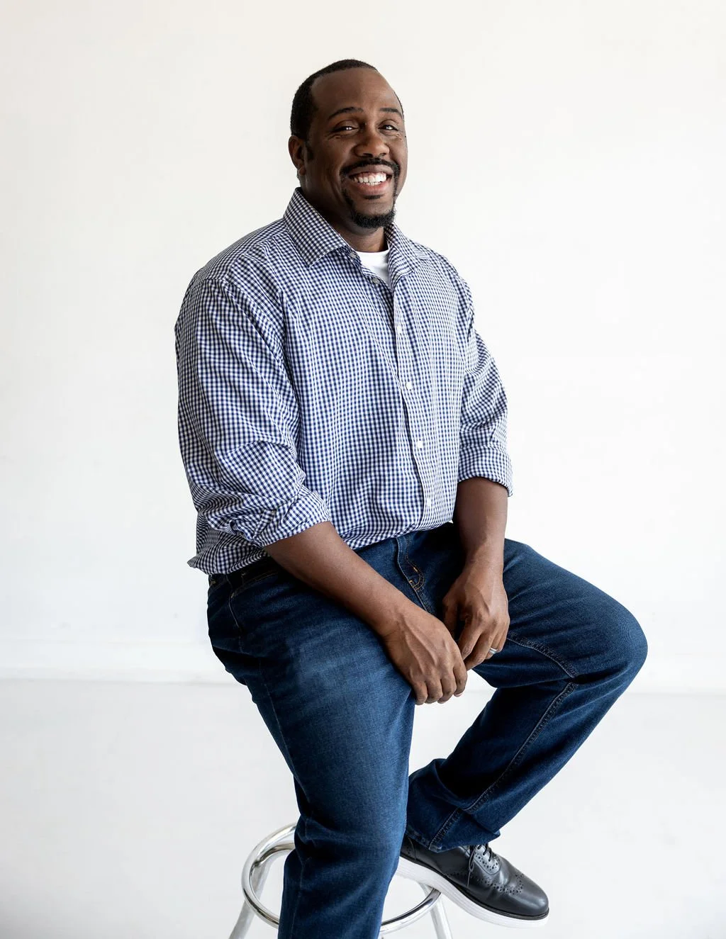 A smiling man with a goatee and short hair, wearing a checkered shirt and jeans, sitting on a stool against a plain white background.