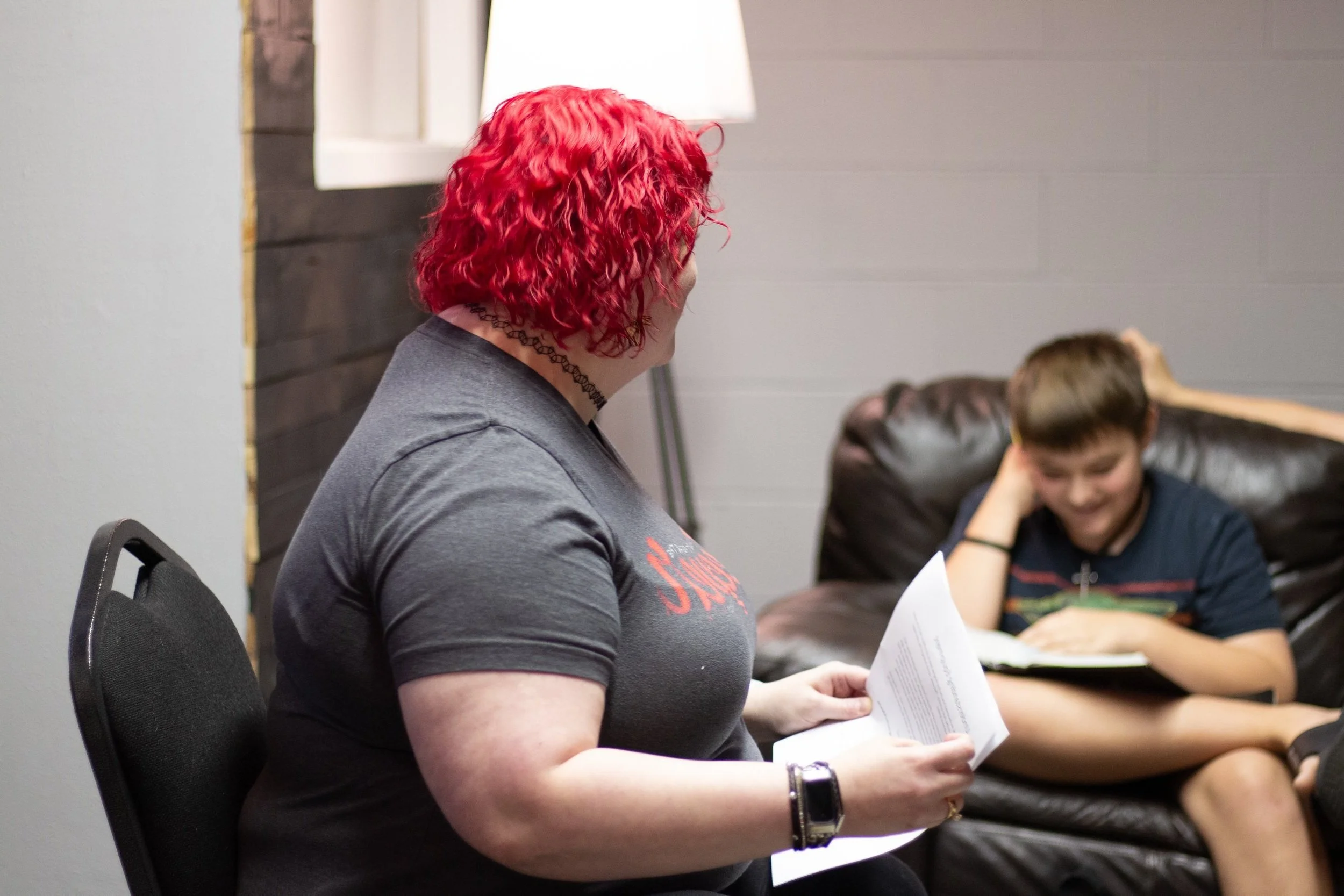 A woman with bright red curly hair and a choker necklace reading a paper to a boy sitting on a brown leather couch, who is smiling and looking down at a notebook.