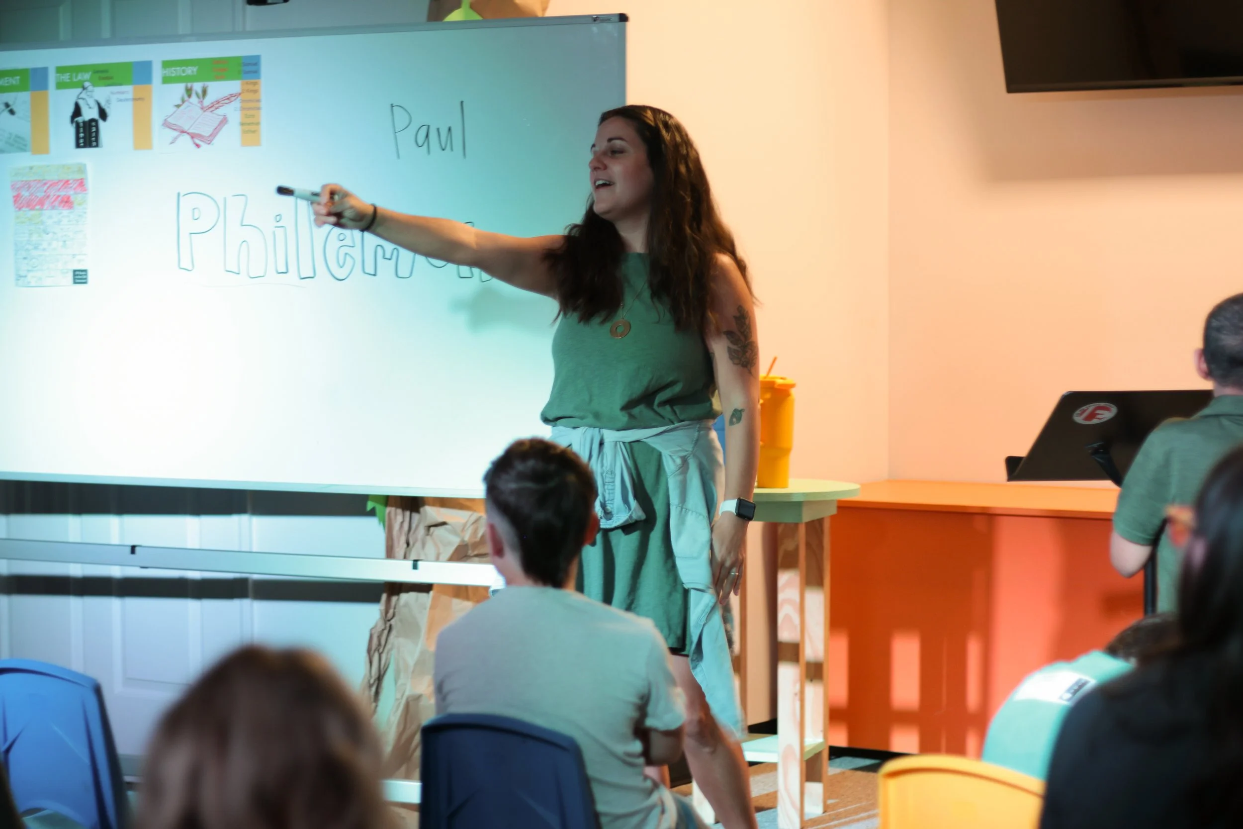 A woman giving a classroom presentation to students, standing at a whiteboard with writing and pictures, pointing at the whiteboard with a marker.