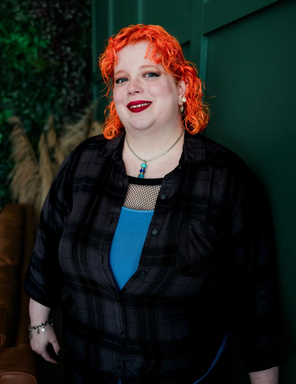 A woman with curly bright orange hair, wearing red lipstick, earrings, a necklace with a blue pendant, a black plaid shirt, and a blue top underneath, standing against a green wall.