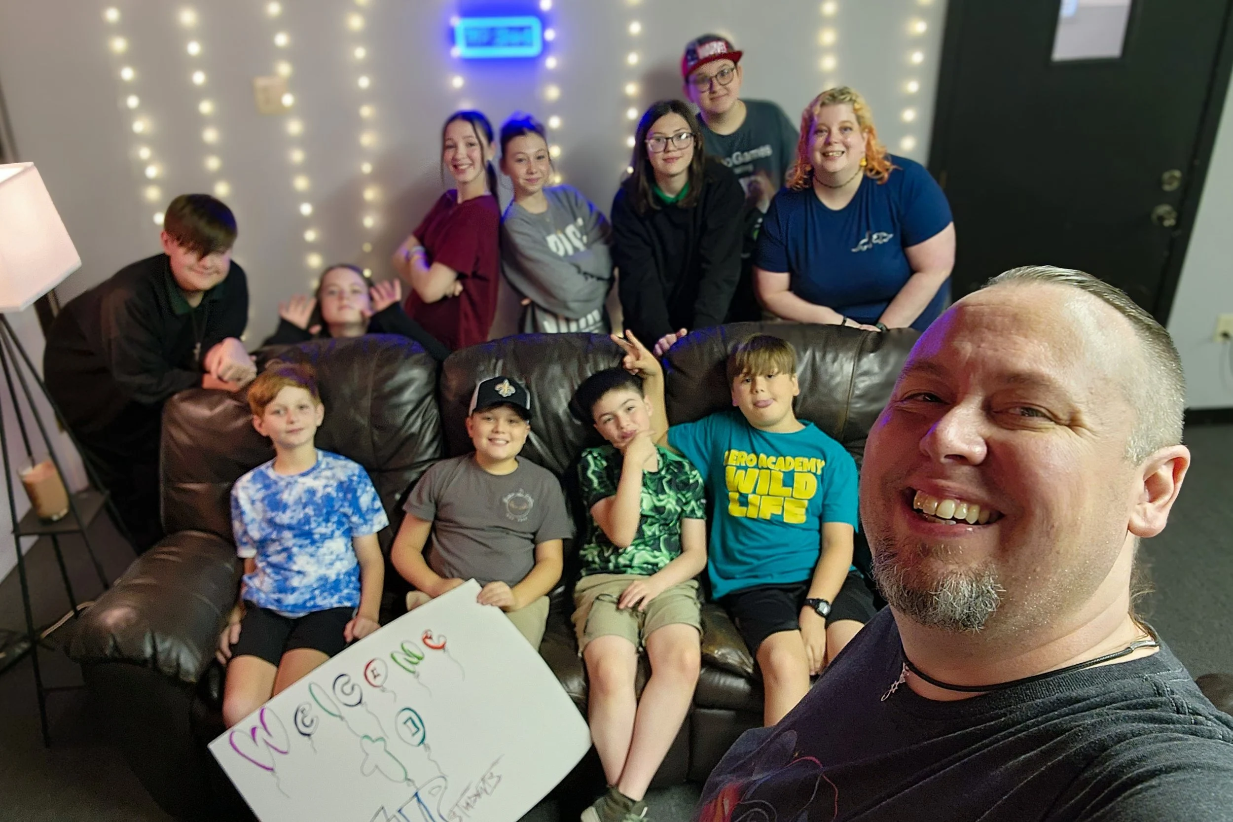 A group of children and adults smiling and posing for a selfie in a room with strings of lights and a blue digital clock on the wall. One child is holding a colorful sign that says "WELCOME."