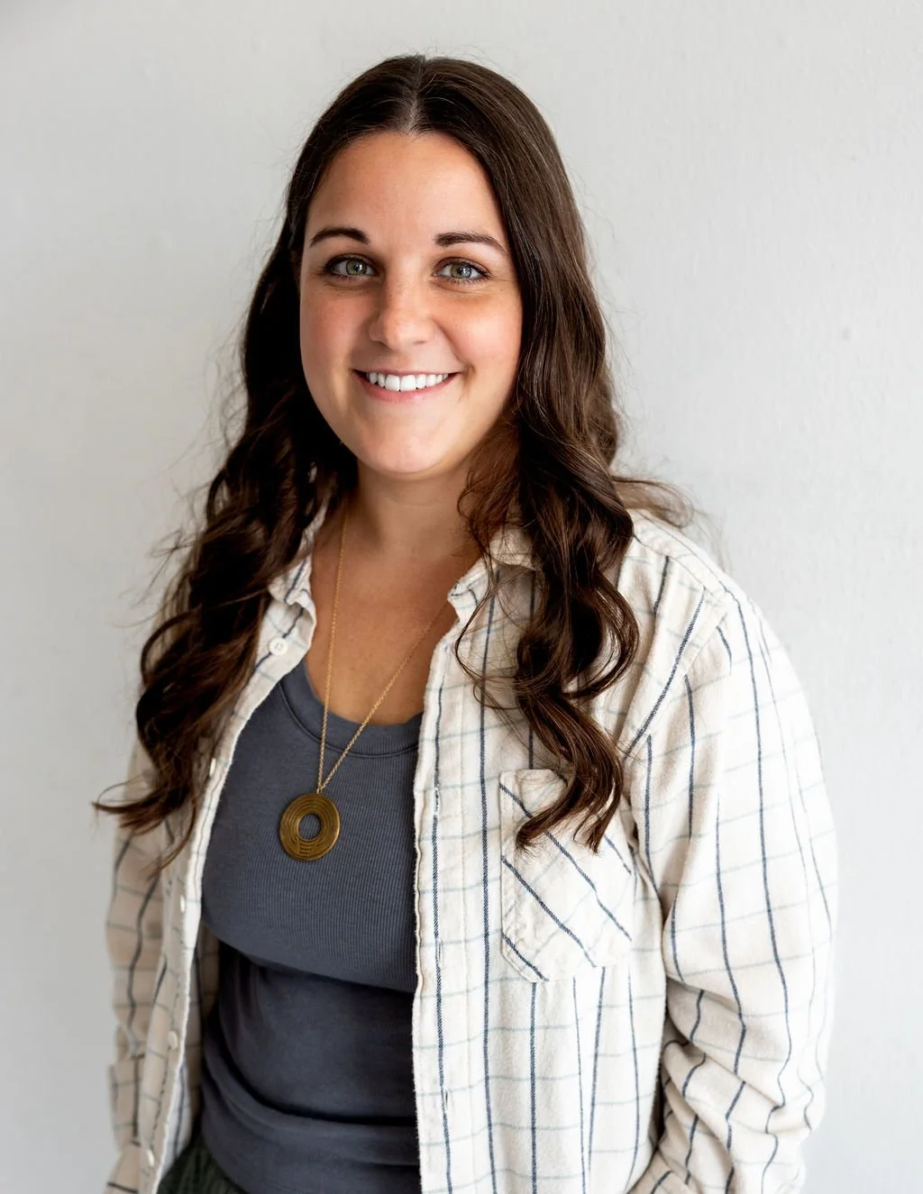 A woman with long brown hair, wearing a dotted shirt over a dark gray t-shirt, and a pendant necklace, smiling at the camera, standing against a plain white background.