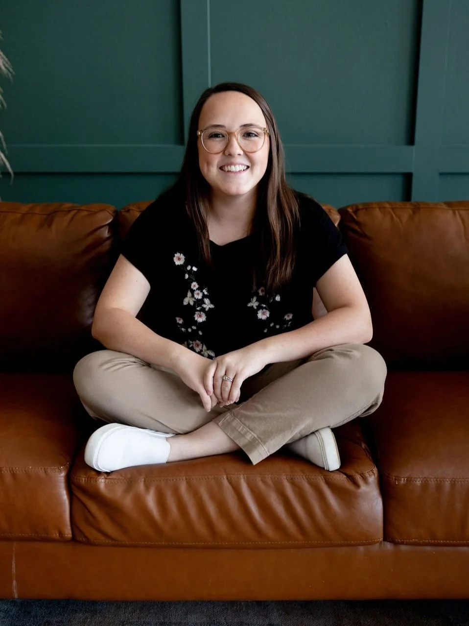 A young woman with glasses, wearing a black shirt with floral embroidery, beige pants, and white shoes. She is sitting cross-legged on a brown leather couch, smiling, in front of a green paneled wall.