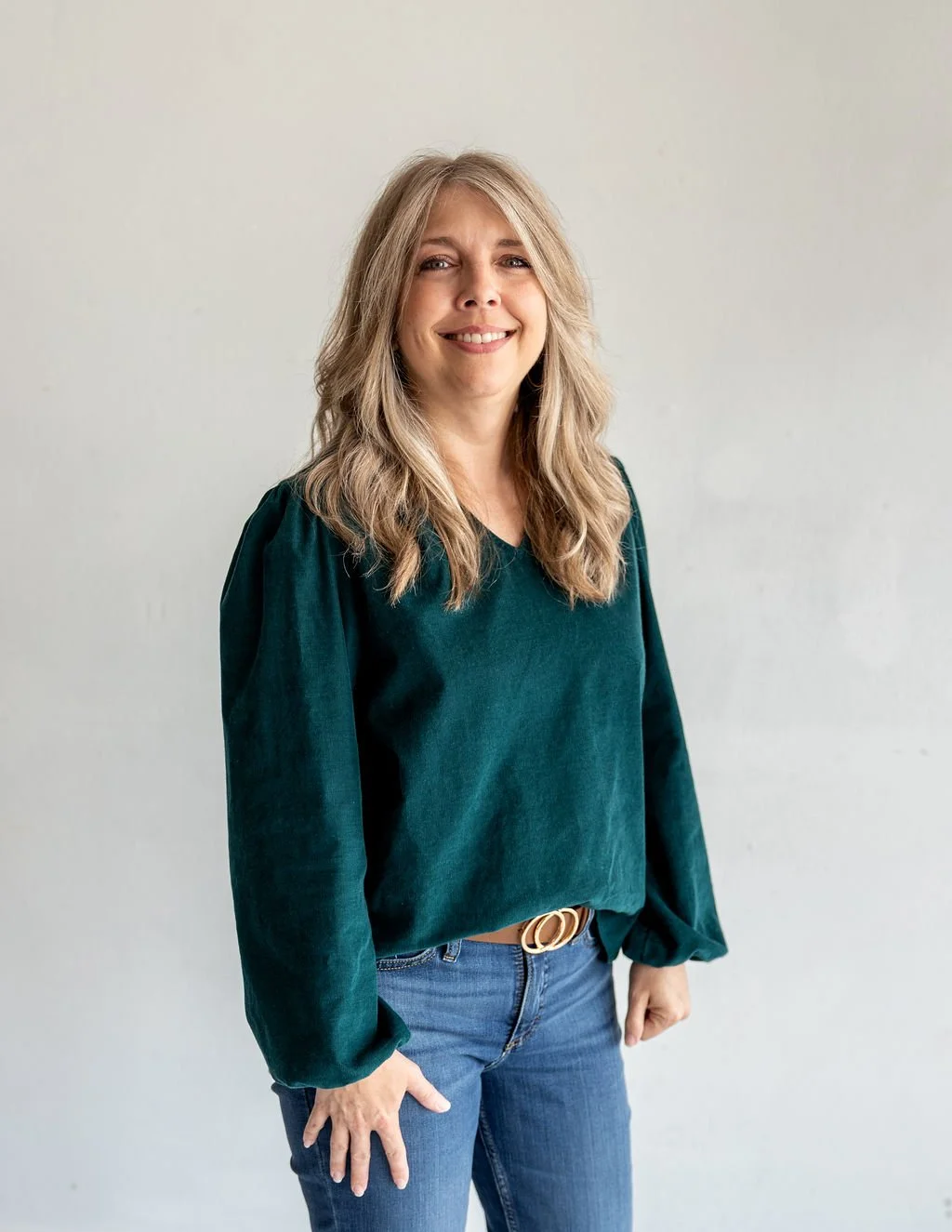 A woman with blonde wavy hair smiling at the camera, wearing a dark green long-sleeve blouse and blue jeans, standing against a plain light-colored background.