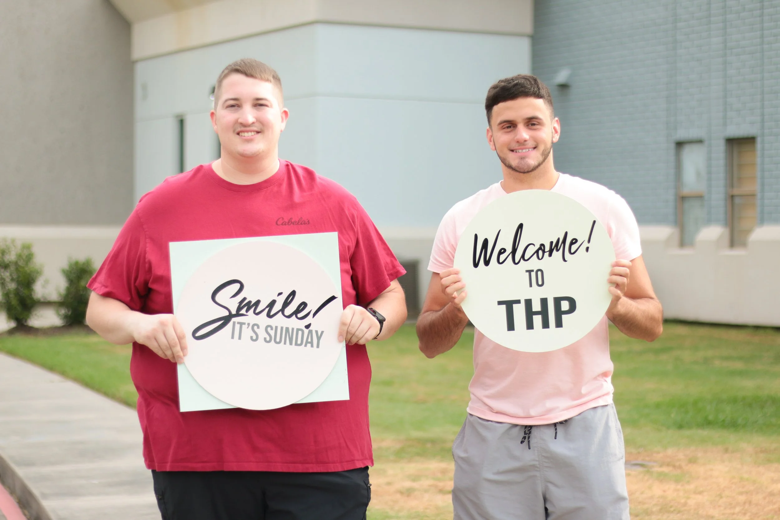 Two young men smiling outdoors, holding signs that say "Smile! It's Sunday" and "Welcome! to THP".