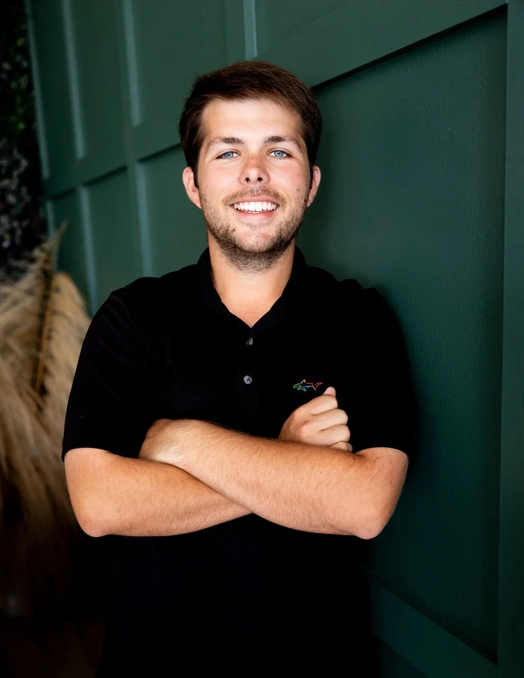 A young man with brown hair and a beard, smiling with arms crossed, wearing a black shirt, standing against a green wall.